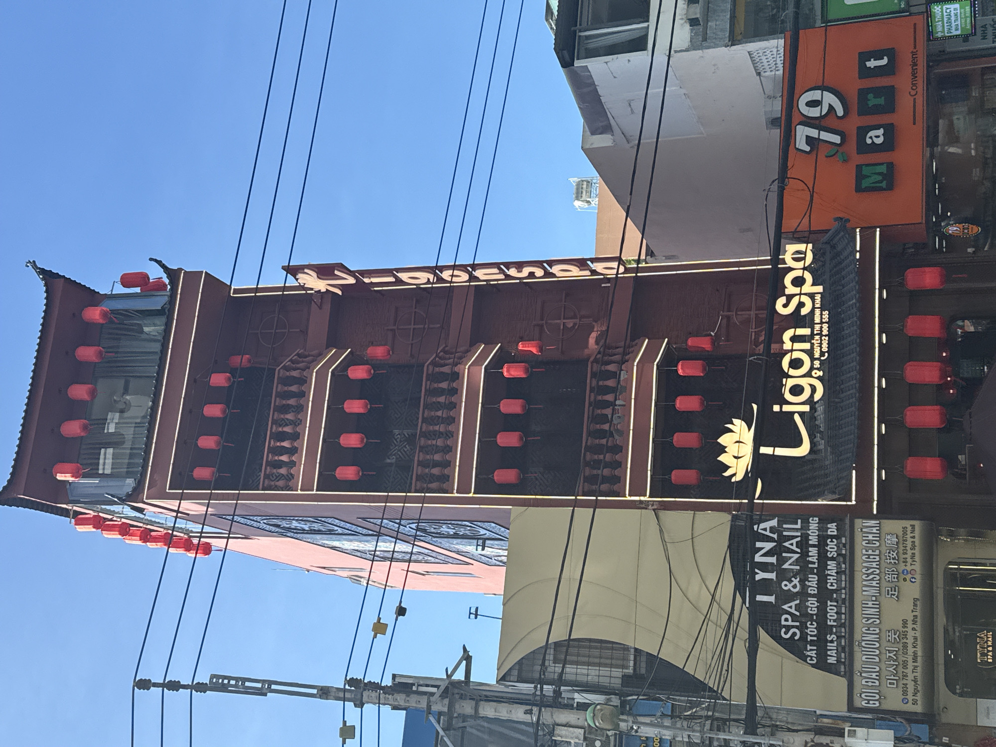 The image depicts a building in Nha Trang, Vietnam, identifiable by signage in Vietnamese. The building's architecture is accented with red lanterns. There are numerous power lines in the foreground. A sign advertises "Ligon Spa," and another adjacent business offers nail and spa services. Another sign advertises a convenience store called "79 Mart." The sky is clear and blue. The scene is an urban street view.