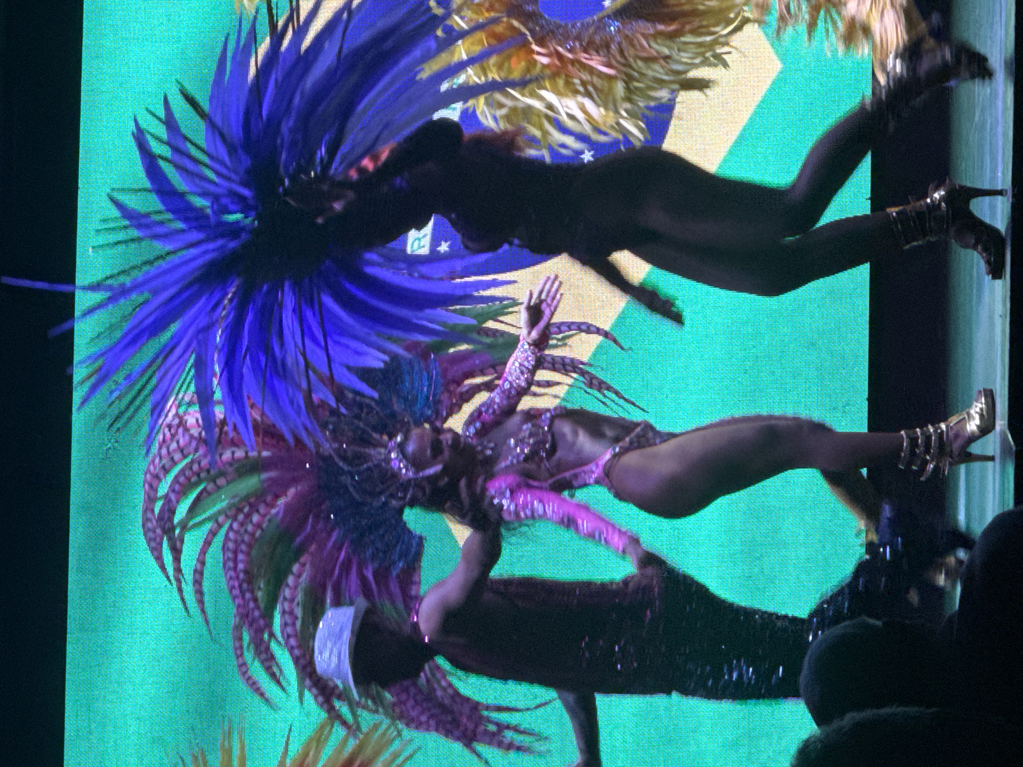 The image depicts a lively performance on a stage, likely in Manavgat, Turkey, given the context provided. Three performers are visible, adorned in elaborate feathered costumes characteristic of a carnival or samba performance. The backdrop is a vibrant green screen featuring a large Brazilian flag.
On the left, a male performer, possibly a dancer or presenter, wears a dark, sequined outfit and a white, sparkling hat. He is shown from the torso up, with his left arm extended, seemingly interacting with the central performer.
The central performer is a woman dressed in a dazzling pink, sequined bikini-style costume with matching feathered headdress and arm adornments. She is captured mid-dance, with her body turned to the right and her right arm extended. She wears elaborate gold platform heels.
To the right, another performer, dressed in dark attire that covers her from head to toe, is also captured in motion. She is partially obscured by large blue and purple feathers from her headdress, which are prominent in the upper right portion of the image. Her legs are visible, and she wears black boots with straps and buckles.
The stage itself appears to be a dark, reflective surface. In the foreground, the silhouetted heads of an audience are visible, suggesting a live event. The overall atmosphere is one of energy and celebration.