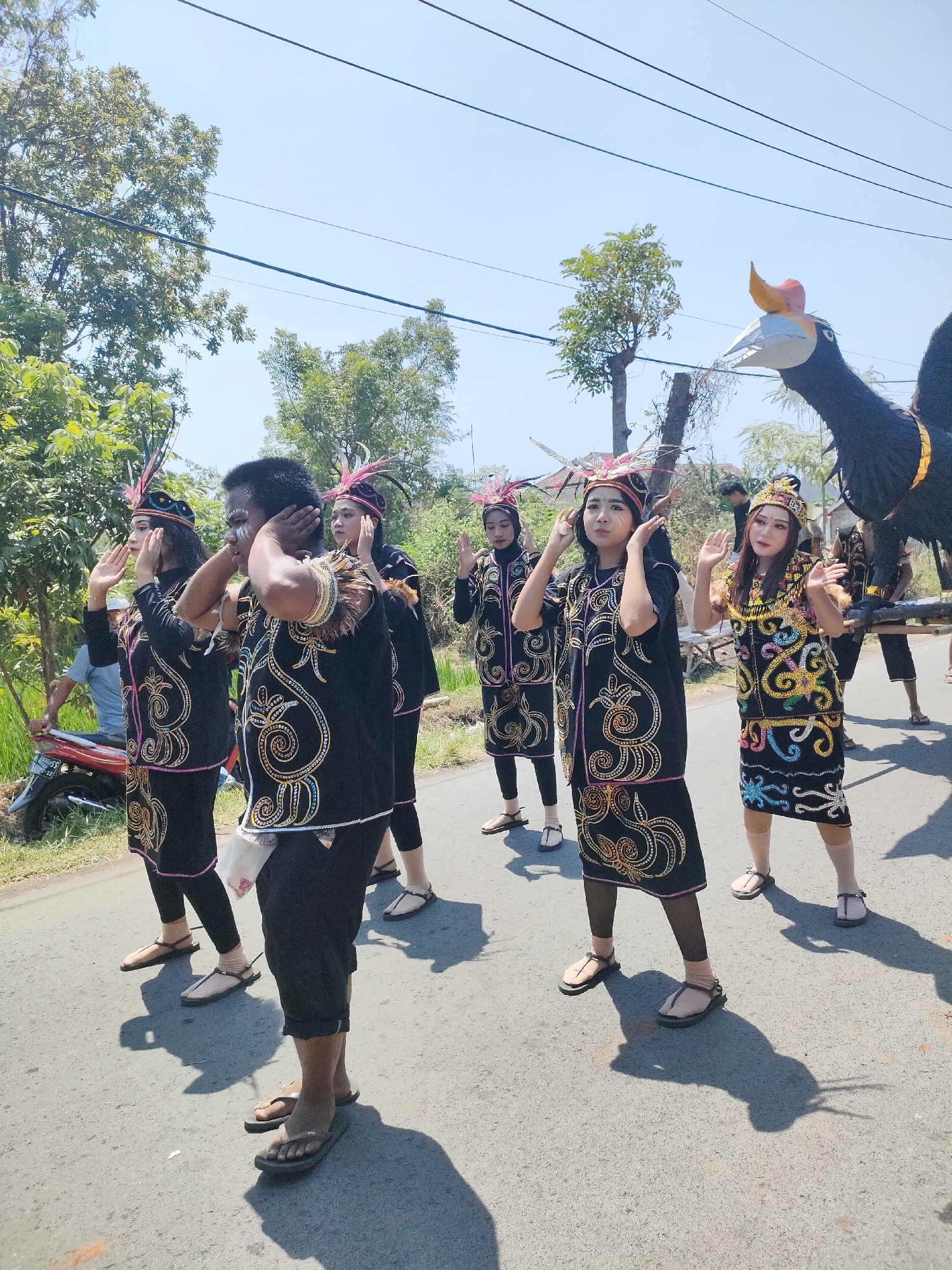 In Tongaswetan, Indonesia, a group of individuals in traditional black attire with ornate gold and colorful embroidery are participating in a procession. The clothing features intricate swirling patterns and feather adornments on their headwear. They are positioned on an asphalt road, with lush green trees and vegetation lining the sides. The sky above is clear and blue, indicating a sunny day. In the background, a large, stylized effigy of a bird with a black body and a white and orange beak is visible, suggesting a cultural festival or parade. Some onlookers are present in the background, and a red motorcycle is parked on the side of the road. The participants appear to be performing a synchronized dance or movement, with their hands raised near their faces.
