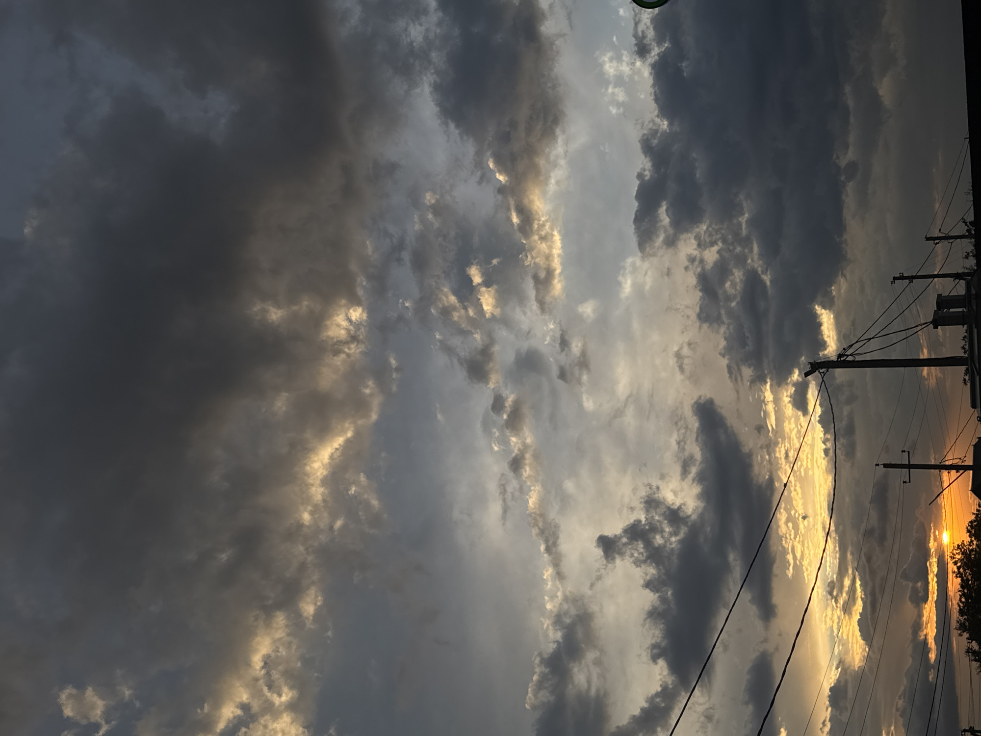The image depicts a stormy sky over Mikhaylovsk, Russia, at sunset. Dark, heavy clouds dominate the upper portion of the frame, with pockets of golden light breaking through from the setting sun near the horizon. The lower third of the image shows silhouetted utility poles and wires stretching across the scene, indicative of an urban or suburban environment. The sun itself is a small, bright disc low in the sky, casting an orange glow on the clouds. No people or specific actions are visible. The overall mood is dramatic and somber, characteristic of impending or recent rain.
