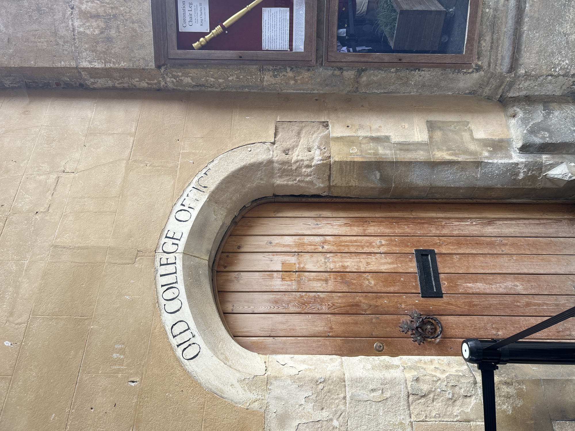 The image shows the entrance to the Old College Office, located at Trinity College, Cambridge, United Kingdom. The entrance features a wooden door with a black mail slot and an ornate, metal door knocker. Above the arched doorway, the words "OLD COLLEGE OFFICE" are inscribed in black lettering on a light-colored stone. To the right of the door, a framed display case is mounted on the wall. This case contains a document and a golden ceremonial staff, with a title that reads "Coronation Chair Leg." The wall itself is constructed of weathered stone blocks. A black stanchion with a retractable barrier is positioned to the left of the doorway, suggesting restricted access. The overall scene depicts an old and historic building entrance.