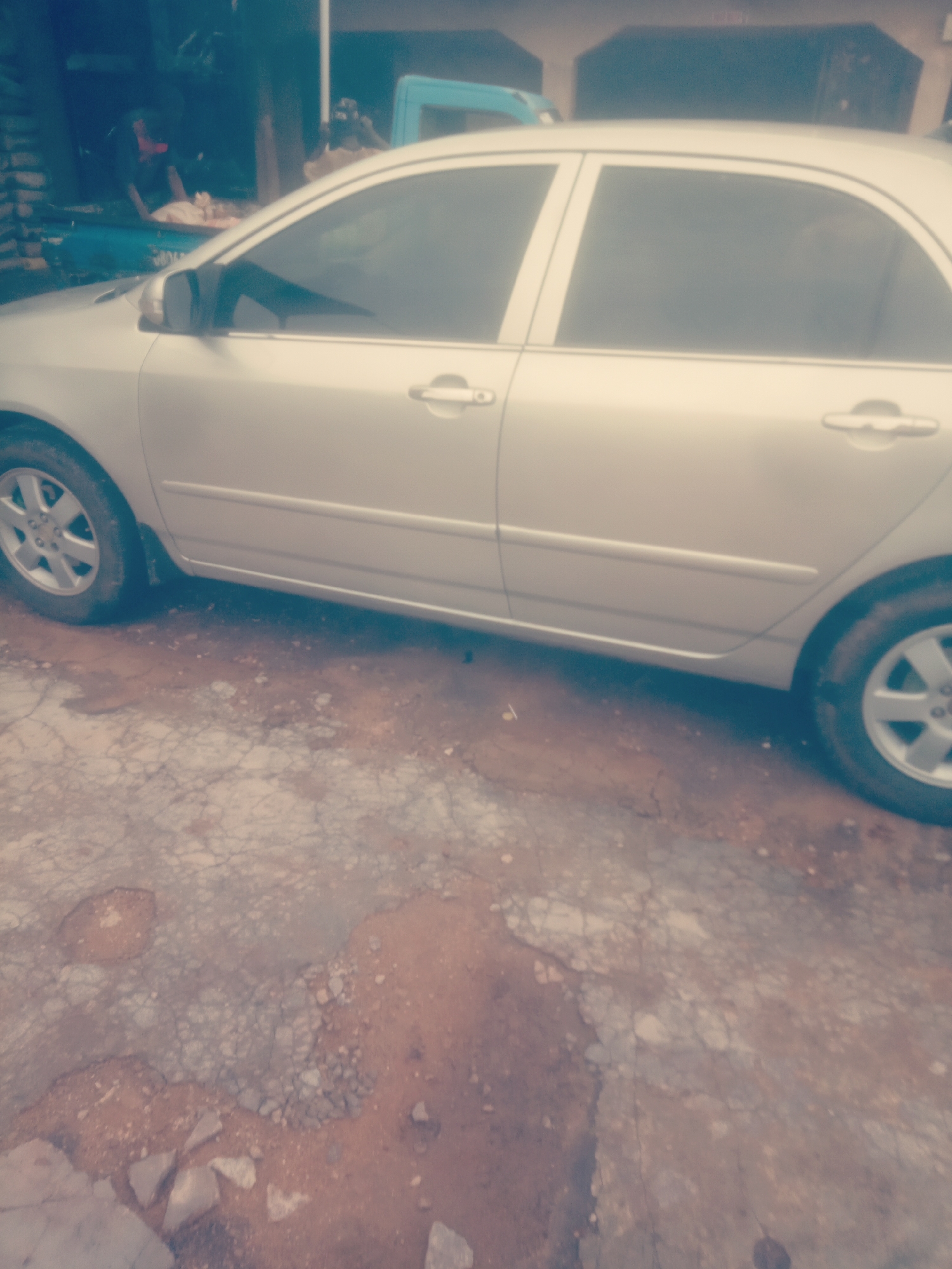 A silver sedan is parked on a dirt and cracked asphalt surface in front of a building. The car's doors are closed and the windows are tinted. The background is blurry and shows a person sitting in a blue vehicle, and some construction materials. The overall image quality is poor due to blurriness and low resolution.