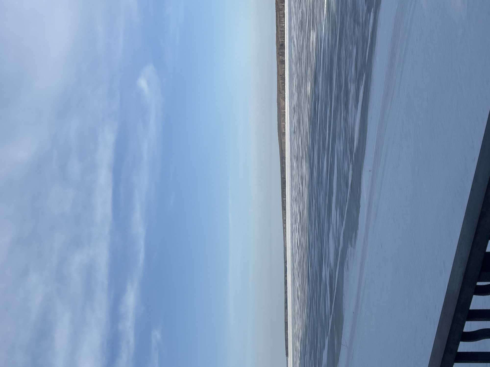 The image depicts a frozen body of water, possibly a lake or river, under a blue sky with wispy clouds. The water is largely covered in ice, with patterns and textures suggesting varying thicknesses and potential cracks. A thin line of trees is visible on the opposite shore. A section of a bridge or railing appears in the bottom right corner, indicating a viewing point. The location is likely in or near the city of Chaikovsky, Russia, given the prompt. The scene is static and provides no direct evidence of people, actions, or interactions.