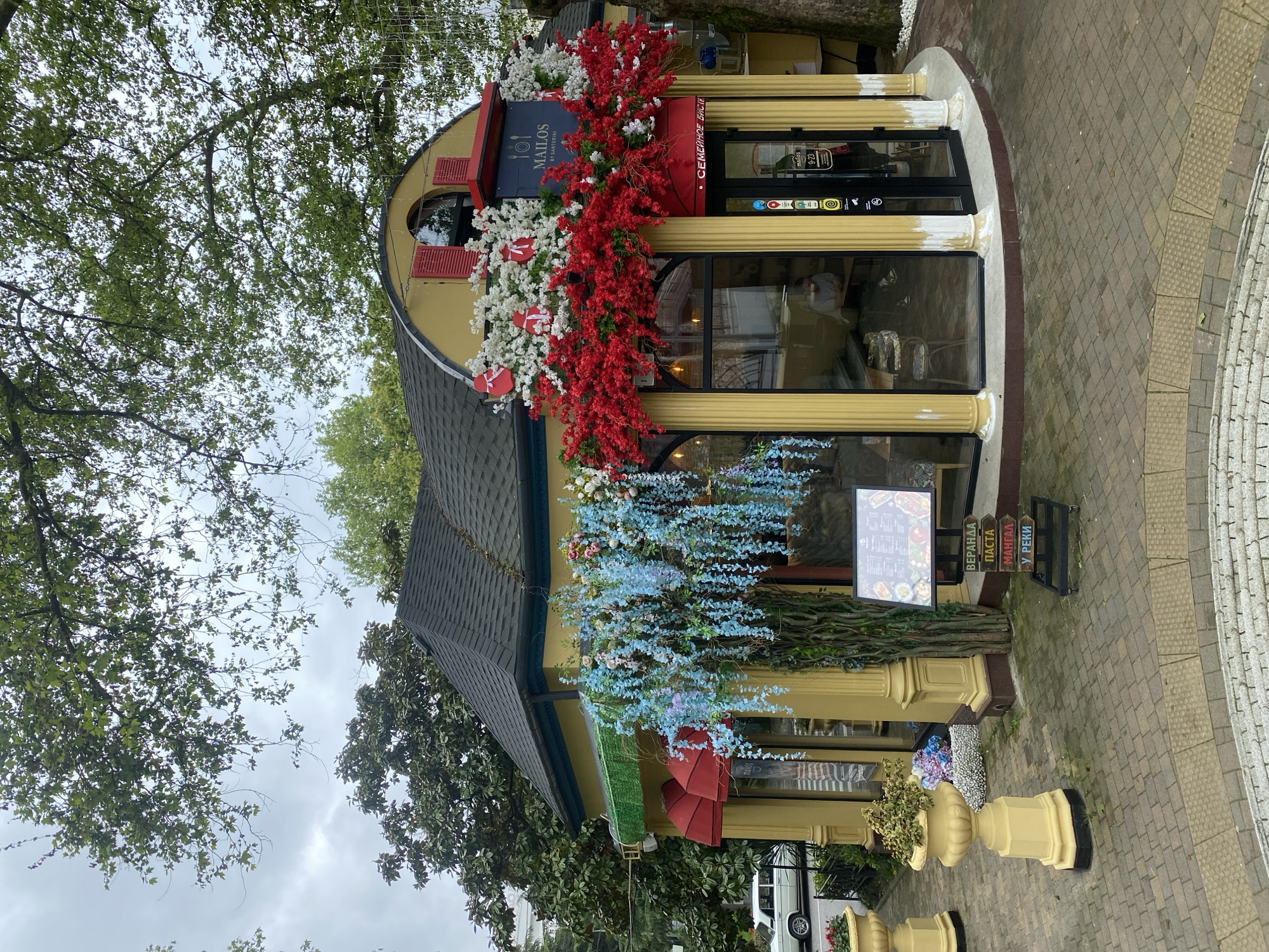 The image depicts the exterior of "Mailos by Santorini," a restaurant located in Sochi, Russia. The building has a unique architectural design with a rounded facade and a pitched roof. The exterior is adorned with vibrant floral arrangements in red, white, and blue. Signage indicates the restaurant's name and menu options. The surrounding environment includes trees, a paved walkway, and street elements. A white car is partially visible in the background.