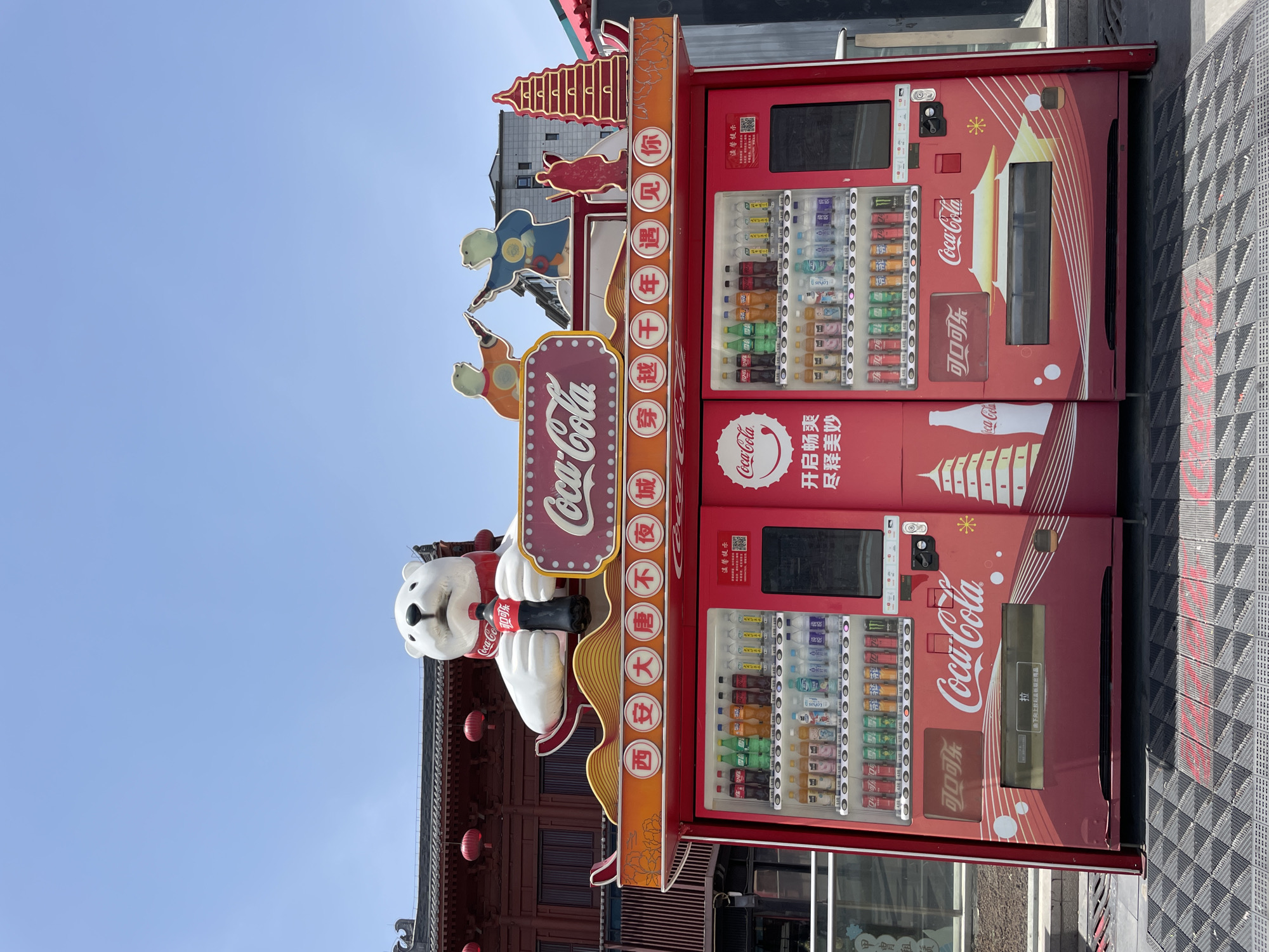 This is a photo of a Coca-Cola vending machine located in the city of Yanta District, China. The vending machine is red and has two sections with different drinks displayed in the dispensers. The machine is decorated with Coca-Cola branding, including a large sign with the Coca-Cola logo, a polar bear holding a bottle of Coca-Cola, and Chinese characters. The machine is also adorned with traditional Chinese designs, such as a pagoda and figures in traditional clothing. The vending machine is located on a street with patterned grey pavement tiles and buildings in the background, including one with red lanterns hanging from the eaves. The sky is visible in the upper left corner and is a clear blue.
