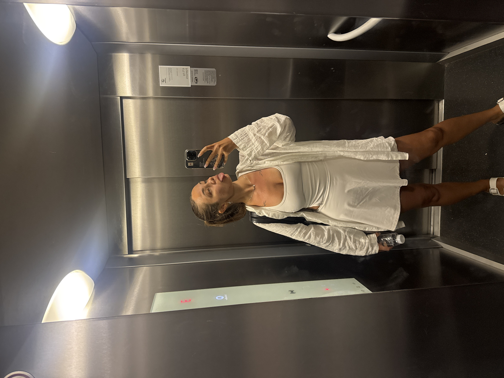 In an elevator with brushed metal walls, a woman with her hair in a ponytail and wearing a white tank top and skirt, and an open white button-down shirt, takes a selfie with her phone. She is sticking her tongue out and has a bottle of water in her left hand. She is wearing white sandals. An illuminated panel with floor numbers is visible to her left, and a white handrail is present on the right wall. The elevator appears to be located in Budva, Montenegro.