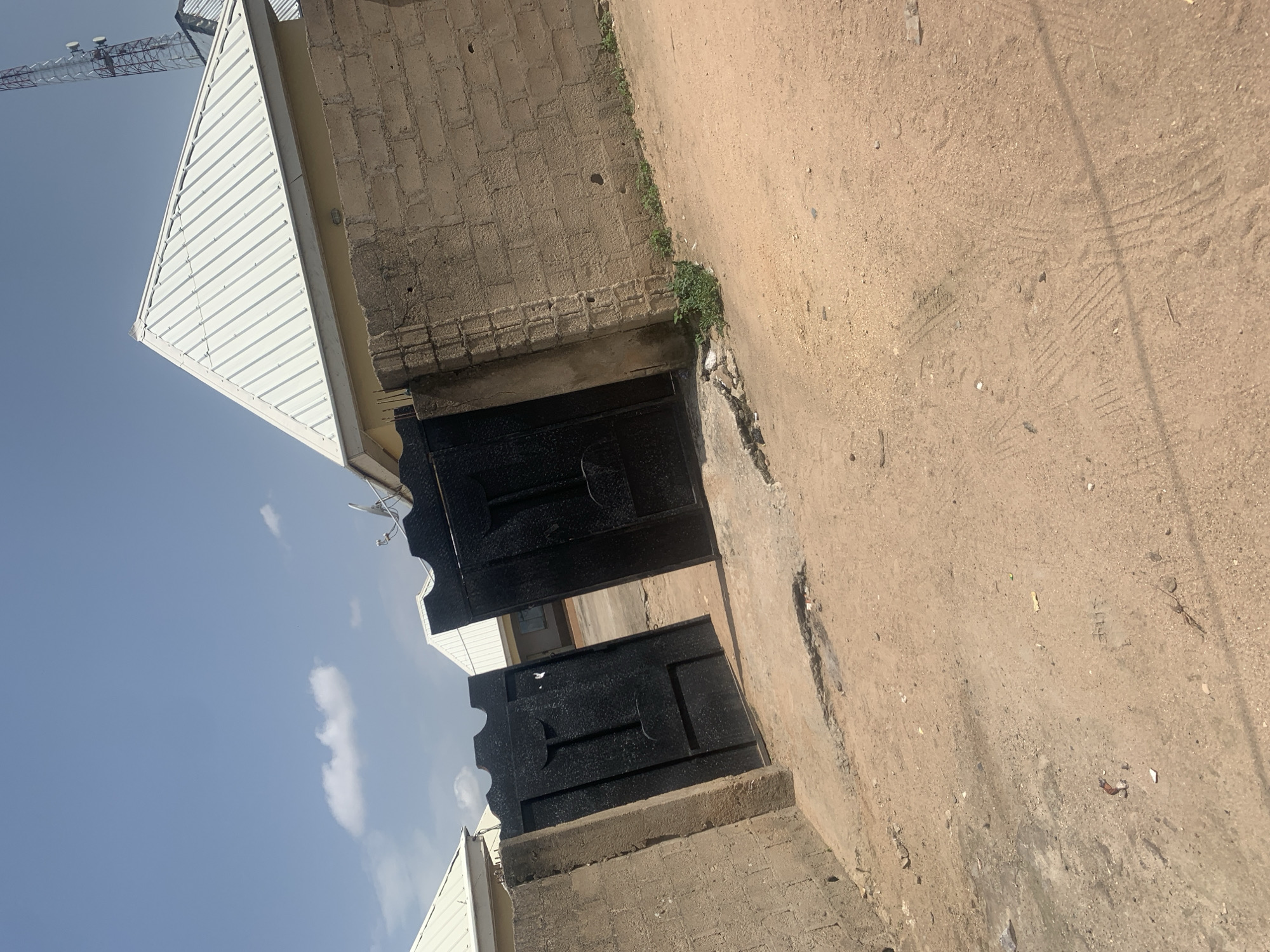 The image depicts an outdoor scene in Jalingo, Nigeria. The primary focus is on a set of closed, black metal gates, one above the other. These gates are set into a wall constructed of concrete blocks. Adjacent to the gates, a building with a corrugated white roof is visible, suggesting a residential or commercial structure. The ground in front of the gates is unpaved and appears to be a dirt or sandy surface, with tire tracks visible, indicating vehicle presence.
In the background, a large communication tower or pylon stands against a clear blue sky. A few scattered white clouds are also present. The lighting suggests it is daytime, likely midday or early afternoon, given the bright sunlight and the shadows cast.
The scene is somewhat unkempt, with small weeds growing near the wall and debris scattered on the ground. The overall impression is of a functional but not elaborately maintained entryway. There is no visible text in the image. The image is oriented vertically, making it appear as though it was taken from a low angle looking upwards.