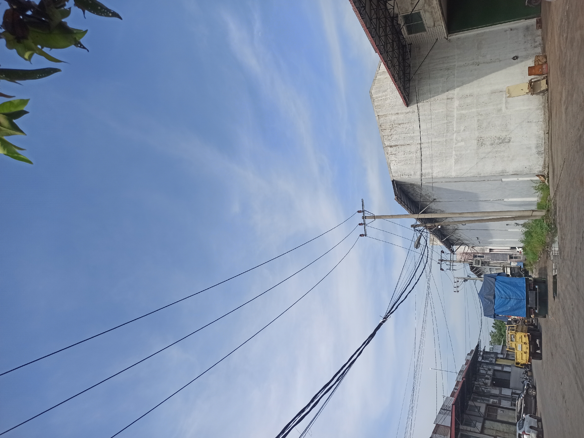 In the city of Medan, Indonesia, a street scene unfolds under a bright blue sky with scattered white clouds. Overhead, a network of thick black power lines stretches across the frame, supported by utility poles. To the left, a row of industrial buildings, some with corrugated metal roofs, line the street. Parked vehicles, including a white SUV and several black cars, are visible in front of these structures. A yellow truck and a larger truck covered with a blue tarp are positioned on the road, suggesting commercial activity. The right side of the street features a large, weathered white building with a dark roof and an attached structure with a red-tiled roof. Near the base of the white building, a metal chair and some barrels are placed. Vegetation, including grass and a few leafy branches in the upper right corner, adds a touch of nature to the urban setting. The ground appears to be a paved surface, with some areas showing signs of moisture.