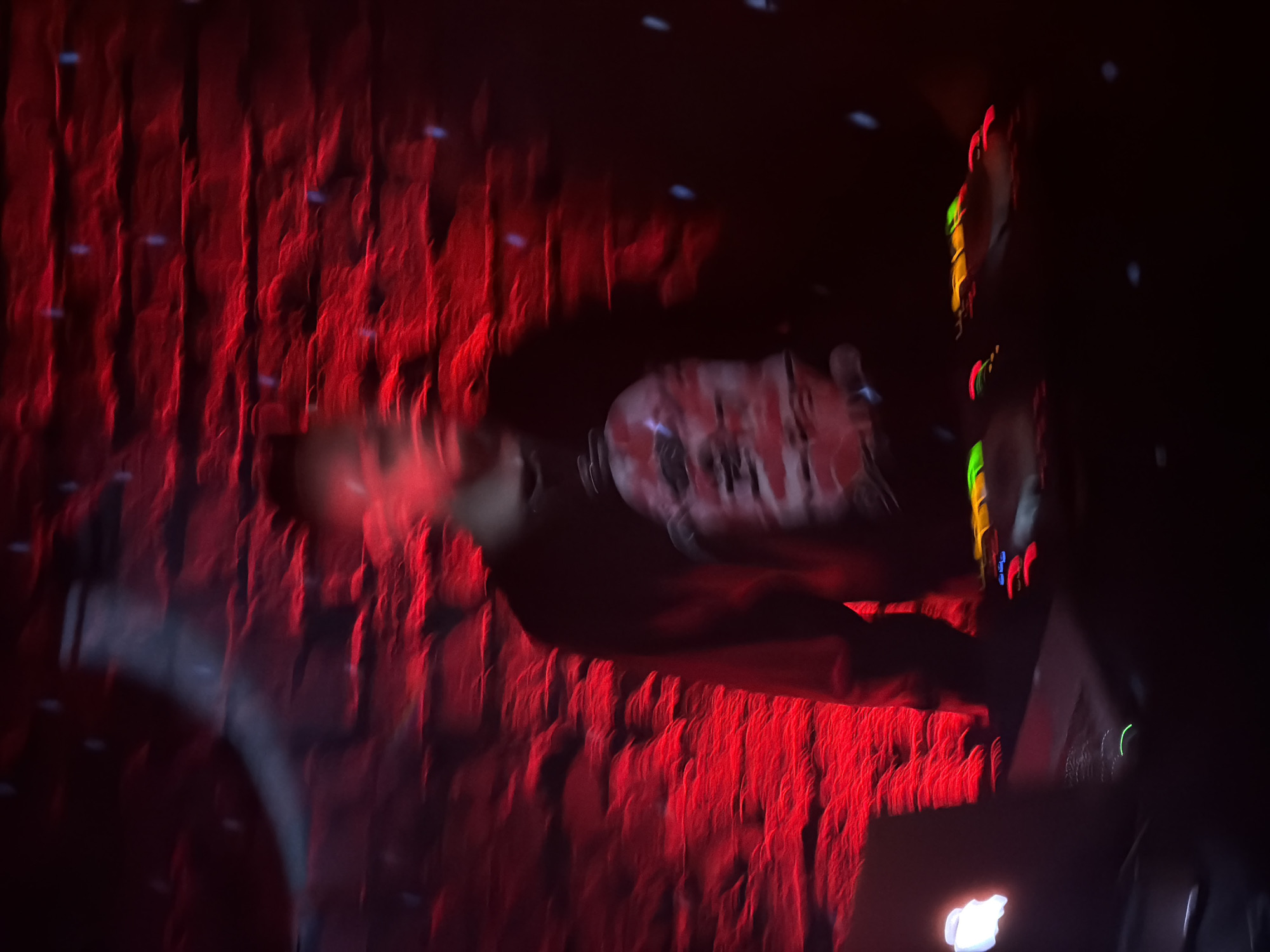 A blurry photo appears to show a person wearing a black shirt with a red and white design, standing on a stage or platform. A red curtain or wall with vertical patterns is in the background. To the right, there is a vertical panel with colorful lights (red, yellow, green). The environment is dimly lit. Given the context of "Moscow, Russia," it is possible this image depicts a concert or performance event held at a venue within the city. However, without further details, this is an educated guess.
