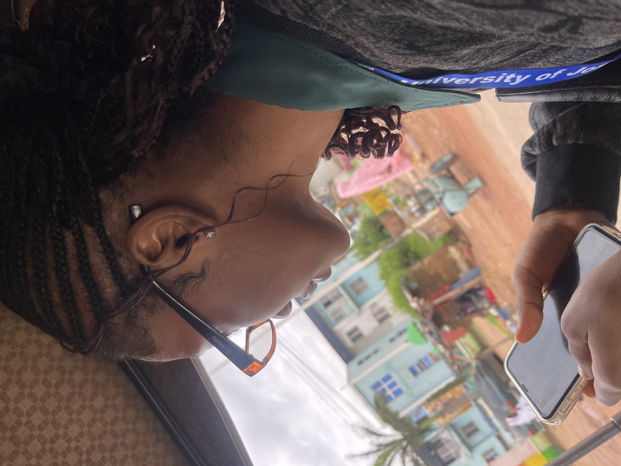 The image shows a young Black woman from the side, wearing glasses and holding a smartphone. She has braided hair, and a lanyard with "University of Jo" visible is around her neck. She is likely in transit, possibly in a motorized rickshaw or similar vehicle, indicated by the blurred background and the interior of the vehicle visible at the top.
The background suggests an urban or semi-urban environment in Rafin Par, Nigeria. Buildings with blue and white facades are visible, along with a palm tree and some rudimentary structures. There are also signs of activity, including what appears to be a roadside market with stalls. A green dome is visible on one of the buildings, which could be a mosque. The overall impression is of a busy, possibly developing, area with a mix of modern and traditional elements. The lighting suggests an overcast or cloudy day.