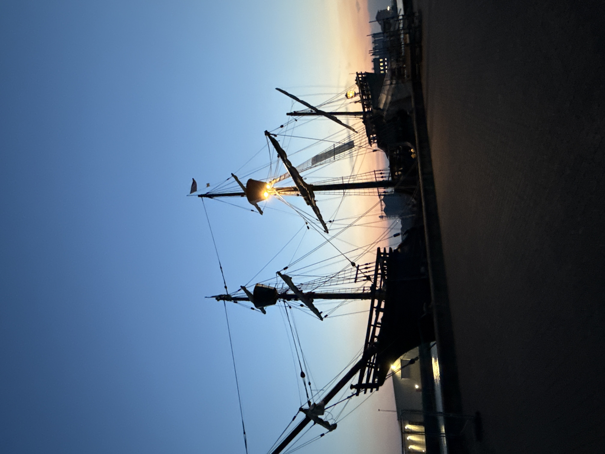 This image depicts a replica of a historical sailing ship docked at twilight in Great Yarmouth, United Kingdom. The sky displays a gradient of deep blue at the top transitioning to soft hues of orange and pink near the horizon. The ship, silhouetted against the twilight sky, is a prominent feature with its tall masts, rigging, and multiple decks. Lights are visible on the ship, illuminating parts of its structure. In the background, a harbor or waterfront area is discernible, with buildings and lights suggesting an urban environment. The foreground shows a paved walkway or ground area, with the overall scene conveying a sense of calm and historical ambiance.
