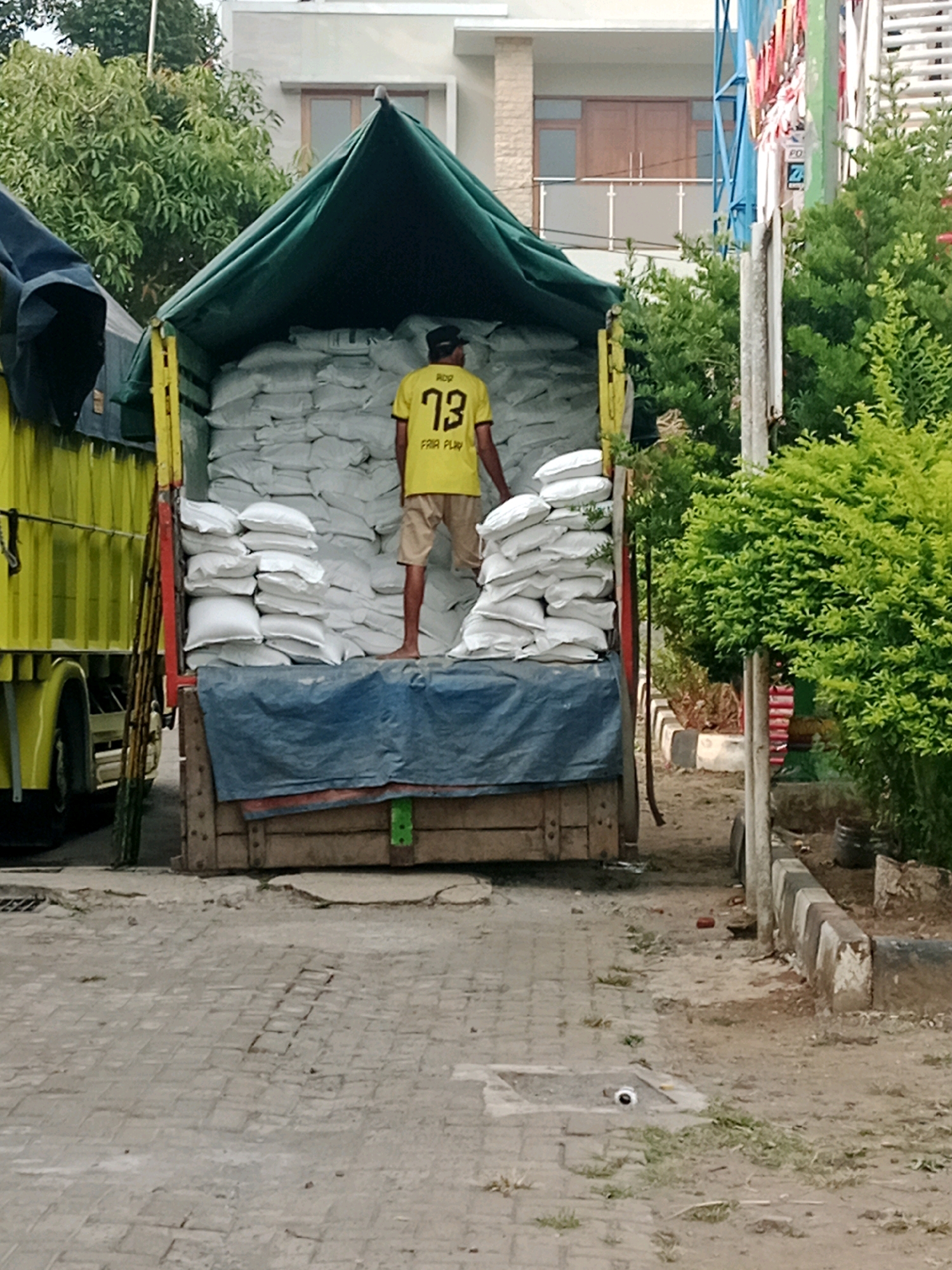 The image depicts a man wearing a yellow t-shirt with the number "73" and the text "FAIR PLAY" on the back, standing on top of a truck filled with white sacks. The truck is partially covered with a green tarp and is parked on a paved surface. Behind the truck, a building with a balcony is visible, along with lush green trees and foliage. The scene suggests a loading or unloading operation in an outdoor urban environment, likely in Salatiga, Indonesia, given the context. The lighting suggests it is daytime.