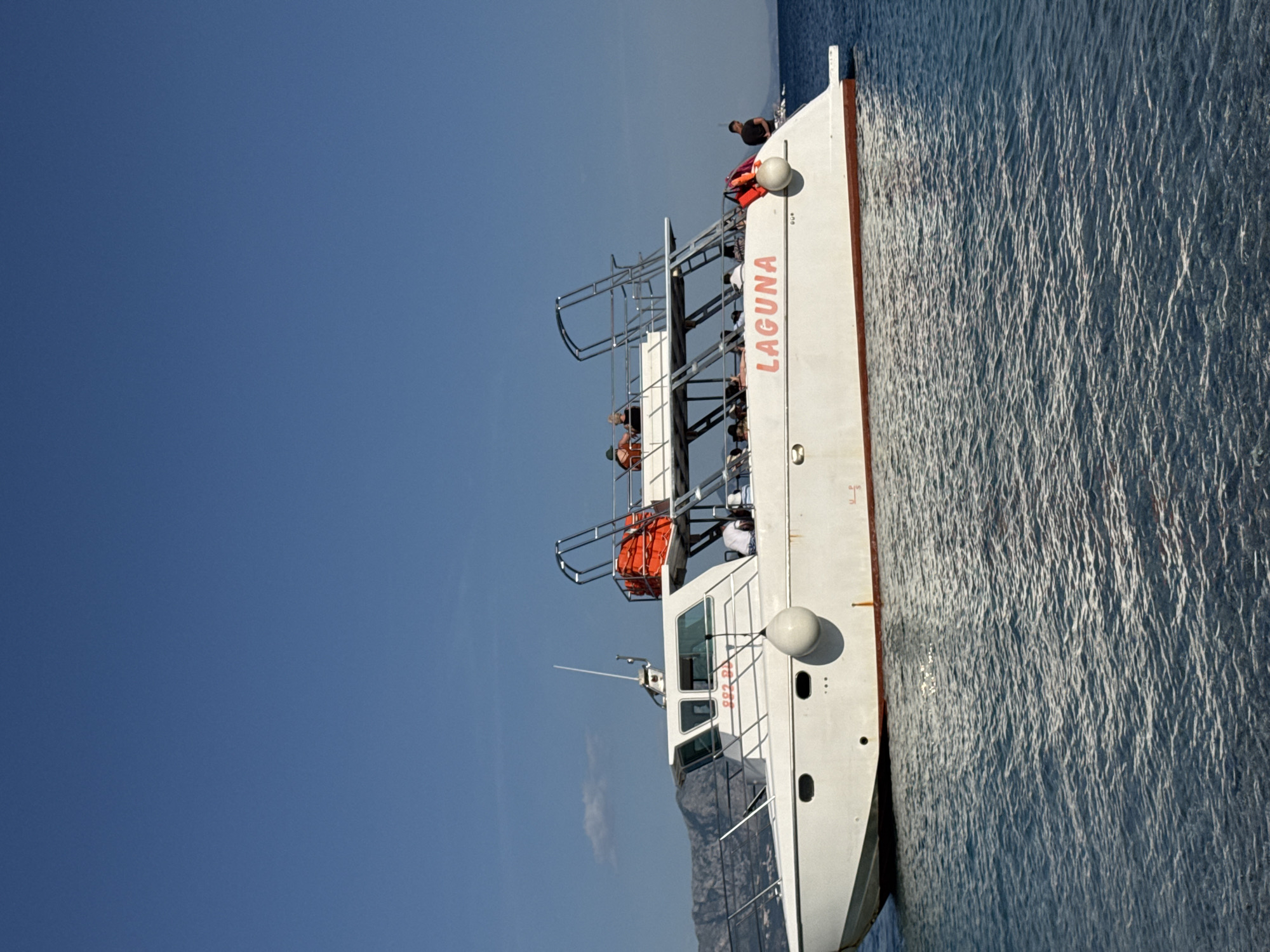 A bright, outdoor shot features the white motorboat "Laguna" adrift on a body of water, likely the Adriatic Sea, set against a clear, light blue sky. The boat's name, "LAGUNA," is painted in red on the hull, near the top of the boat. Passengers are visible on the upper levels of the boat, with one passenger standing on the top deck. The boat is equipped with life rafts or other safety equipment.
In the background, a mountainous landscape is faintly visible on the horizon. The location is likely near Prijevor, Montenegro, which is located on the Adriatic coast. The presence of the boat, the body of water, and the mountainous terrain support this location.
