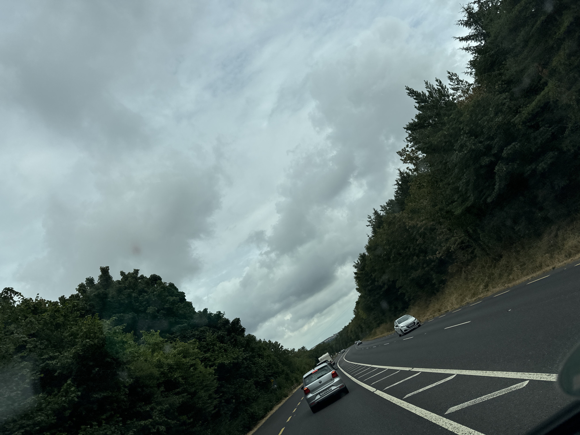 The image depicts a view from a vehicle traveling on a highway in Glenmore, Ireland. The road, marked with white lines and a yellow verge line, curves to the left. Several cars are visible on the road. Dense, dark green trees line both sides of the highway. The sky is overcast with heavy grey clouds. The scene is shot from a low angle, likely from the driver's perspective or a passenger in the front seat.