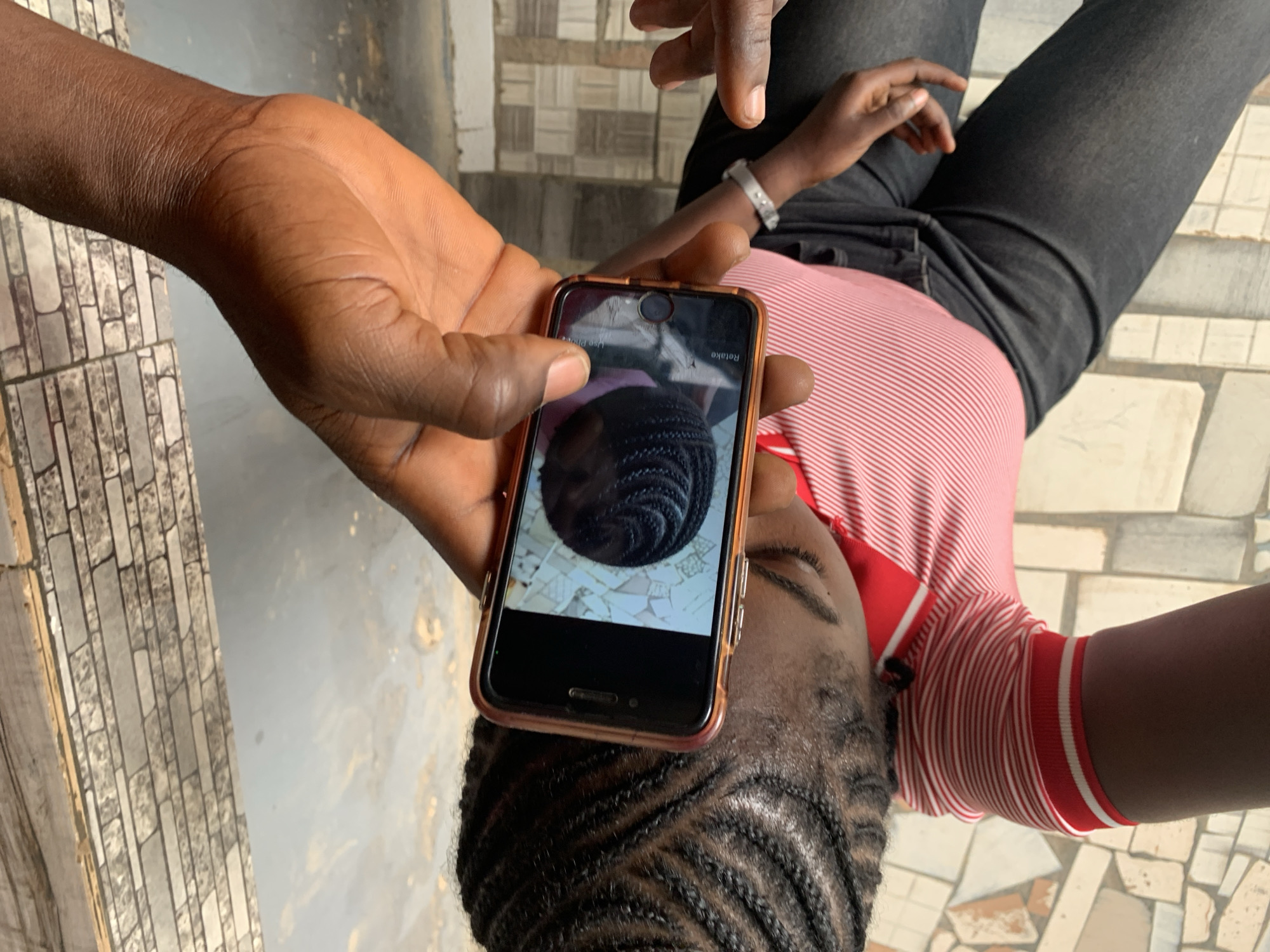 An overhead, slightly angled close-up image shows a person holding a smartphone, presenting its screen to another individual. In the foreground, a dark-skinned hand firmly grasps a smartphone, which is encased in a rose gold or copper-colored protective cover. The phone's screen displays a close-up image of tightly braided, dark hair, possibly a reference picture. Text visible on the phone screen includes "Use Photo" on the left and "Retake" (inverted) on the right, suggesting a photo selection interface. A small crack is visible on the phone screen near the top edge.
Partially visible in the background is another dark-skinned individual, seen from the chest up. They are wearing a red and white striped polo shirt with a red collar and white stripes on the sleeves, paired with dark grey or black jeans. Their hair is styled in neat, tight braids, similar to the image displayed on the phone. A silver-toned watch is worn on their left wrist. Their eyes appear closed or cast downwards, suggesting a moment of contemplation or rest.
The setting appears to be indoors or under a covered area, with a patterned tiled floor or wall visible behind and around the subjects, featuring rectangular shapes in shades of grey and beige. The lighting is bright, indicative of daytime. The scene captures a casual interaction, likely related to hair styling, taking place in New Stadium, Nigeria.