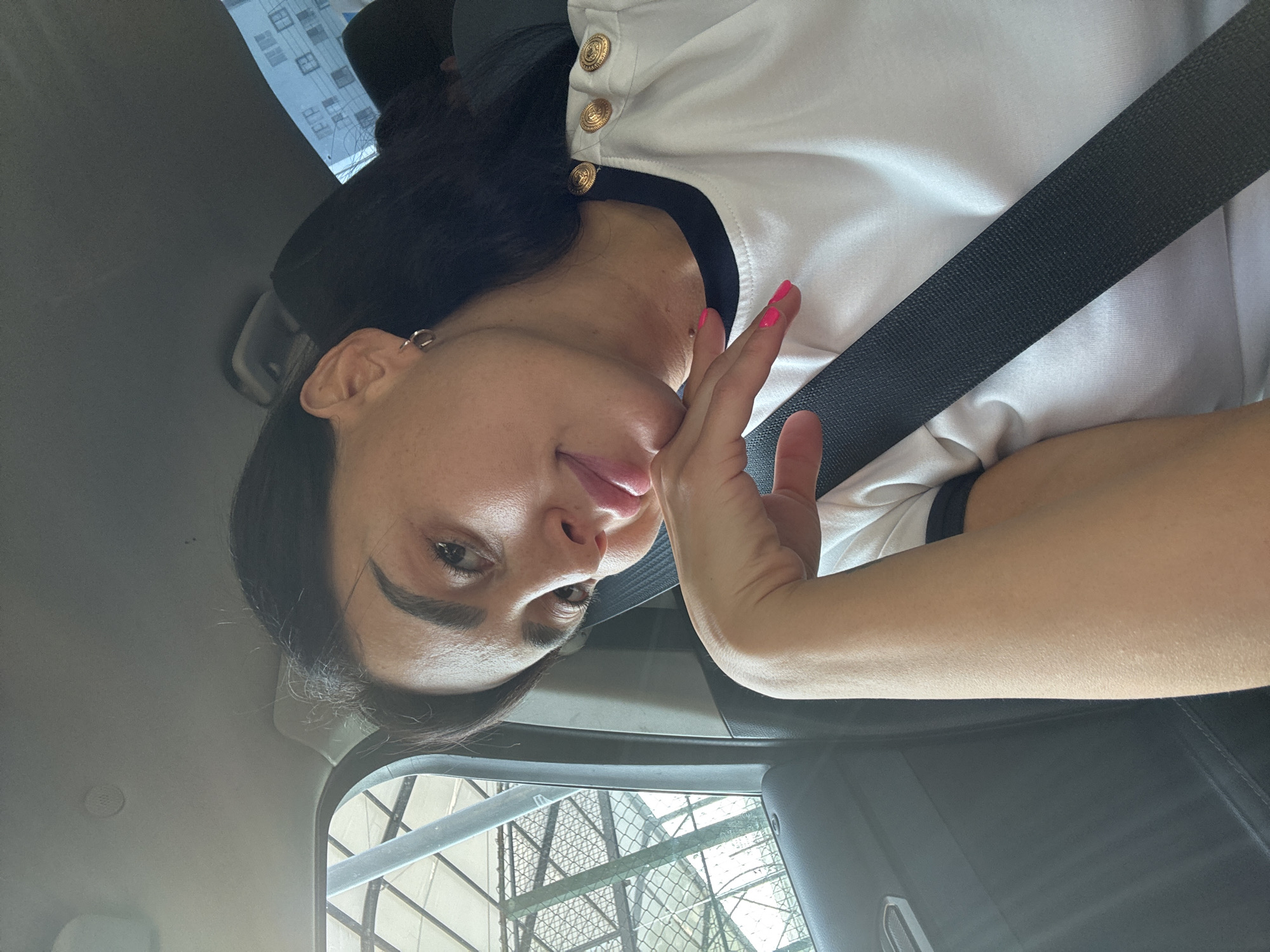 A woman with dark hair and a white shirt with gold buttons is seated in a car, likely in Tbilisi, Georgia, as indicated by the surrounding environment. She is looking towards the camera with one hand resting on her chin and her tongue slightly extended. A black seatbelt is fastened across her chest. Through the car window, a building with multiple windows and what appears to be a metal fence or structure can be seen. The interior of the car is visible, including the dashboard and headliner. The lighting suggests it is daytime, possibly overcast.