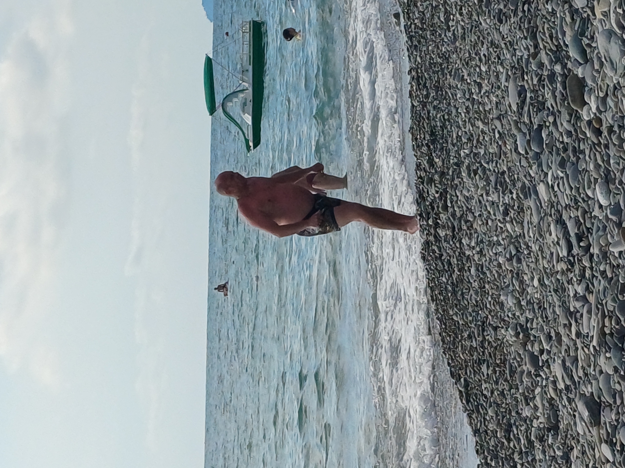 A man with a shaved head and light skin, wearing dark patterned swim trunks, stands on a pebble beach by the sea. He is facing left with his body angled slightly towards the camera, holding his right leg up and bent at the knee, with his right hand on his shin. Gentle waves are breaking on the shore around his feet. In the background, the blue sea extends to the horizon under a pale sky with scattered clouds. A green and white boat with a canopy is on the water to the right of the man, and a person is swimming in the sea near the boat. Further in the distance, another person is visible sitting on a small object in the water. The overall scene appears to be a typical summer day at a beach resort. This is a view of the Black Sea coast in Sochi, Russia.