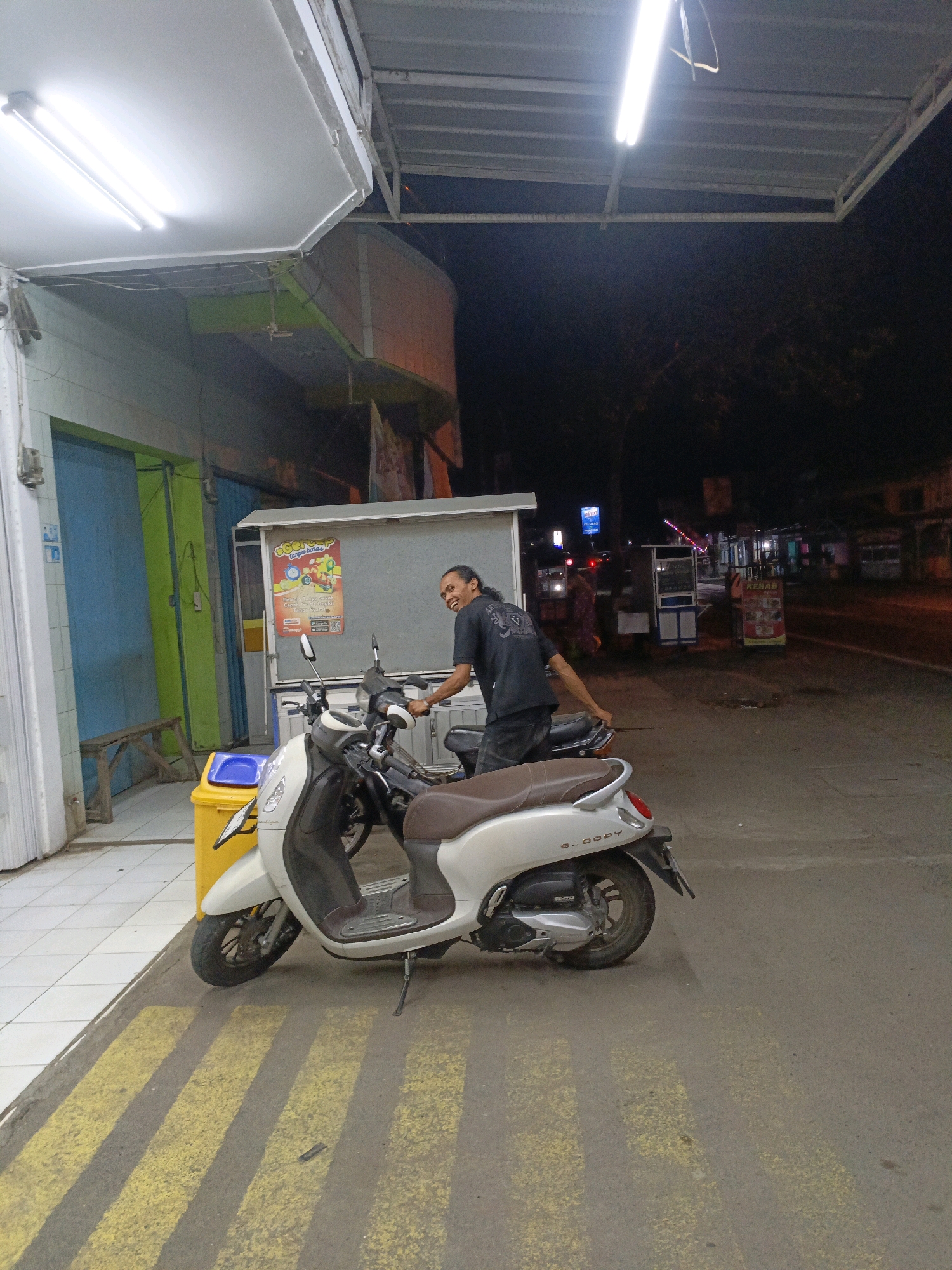 The image captures a street scene at night in Purwajaya, Indonesia. In the foreground, a white Honda Scoopy scooter is parked. Behind it, a man with dark hair is leaning on another scooter, smiling towards the camera. He is wearing a black t-shirt. To the left of the scooters, a yellow trash can with a blue lid is visible. The background reveals a street with several small businesses and a road. The overall lighting suggests it is after sunset, with artificial lights from shops and streetlights illuminating the area.