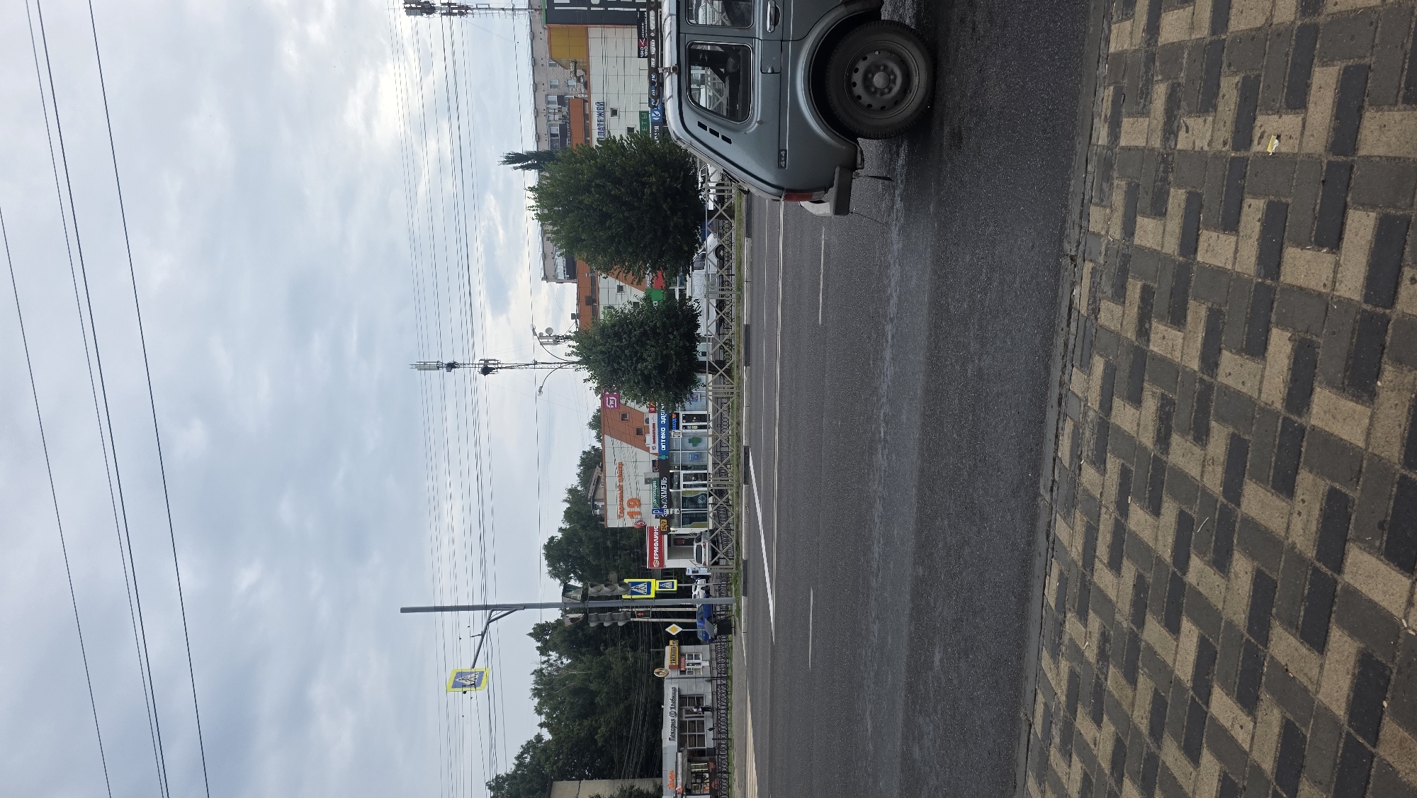 A street view in Tambov, Russia, shows a grey sedan in the foreground on the right, facing left and slightly away from the camera. The car is parked on a wet asphalt road. The road is divided by white lines and a white pedestrian crossing is visible in the middle ground. On the left side of the road, there are several buildings with storefronts, including a bakery and a pharmacy, as well as a traffic light and a pedestrian crossing sign. Trees line the sidewalk in front of the buildings. Overhead, power lines are strung across the frame, and two cell towers are visible in the distance. The sky is overcast with grey clouds. The sidewalk in the immediate foreground is paved with interlocking bricks in a dark grey and beige pattern.