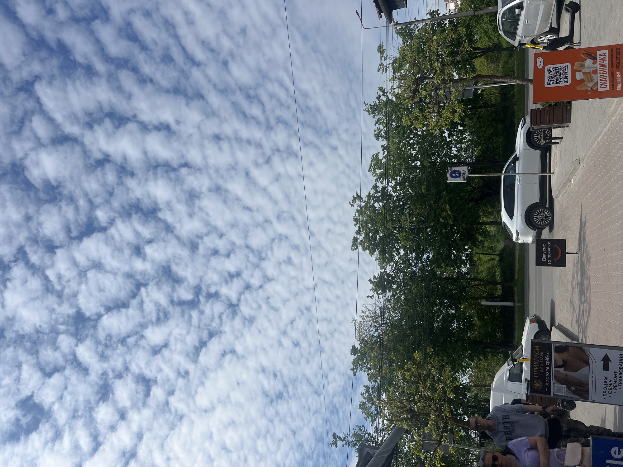 The image depicts a street scene in Ternopil, Ukraine. The sky is filled with altocumulus clouds. Along the sidewalk, several signs are visible, including one for "СКАРБНИЧКА" (Skarbnychka), a bakery offering a loyalty program, and another for "ПРИКРАСИ ДЛЯ ПАНІ" (Jewelry for ladies). There are parked white cars and trees lining the street. People are visible near one of the signs. The OCR text reveals additional information from the signs.