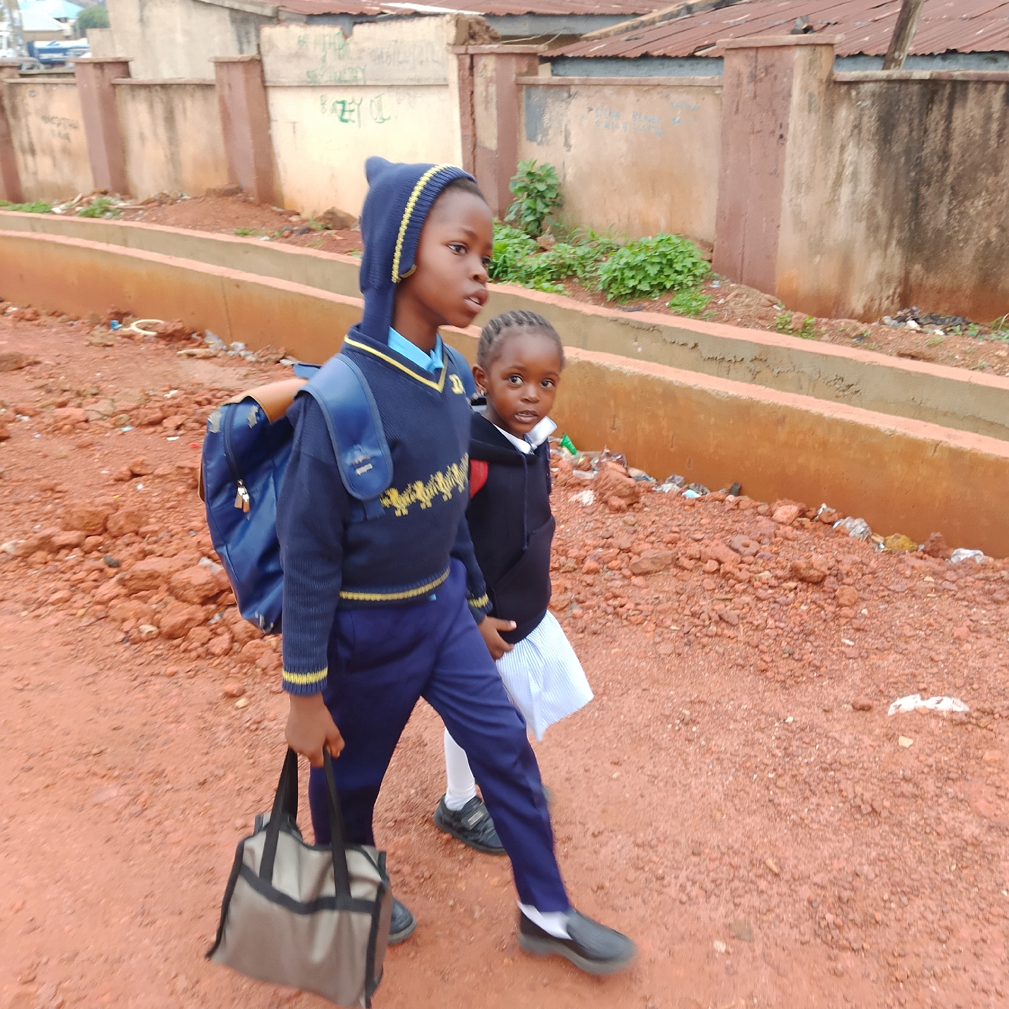 Two young girls, dressed in school uniforms, walk on a reddish-brown dirt road. The older girl, on the left, wears a dark blue sweater with yellow trim and a matching blue knit hood with yellow stripes. She carries a blue backpack and a light gray and black tote bag. The younger girl, to her right, wears a black hooded jacket over a white shirt and a pleated white skirt. She has braided hair. They are walking alongside a raised concrete structure that borders a path. In the background, there are buildings with corrugated metal roofs and faded walls, some with graffiti. Greenery and scattered debris are visible. The environment appears to be an urban or semi-urban setting in Gura Top, Nigeria.