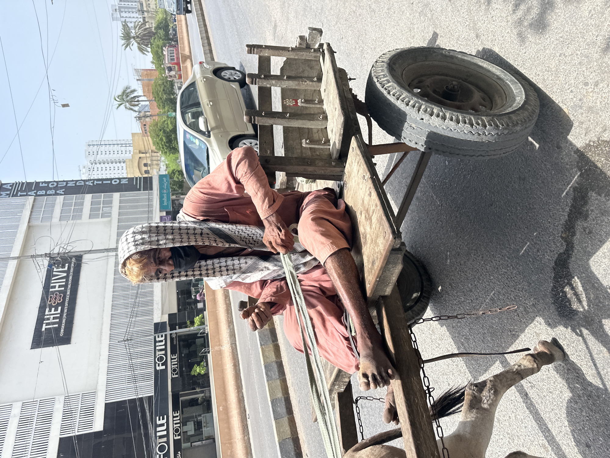 In Karachi Division, Pakistan, a man wearing a headscarf and mask is seated on a wooden cart being pulled by a horse. He is dressed in a salmon-colored shalwar kameez and is holding reins. The cart has two wheels and a wooden structure at the back. Behind the cart, a busy street is visible with cars and buildings. One building in the background has a sign that reads "THE HIVE" and "FOTILE." The scene appears to be a typical urban setting with both traditional and modern elements.