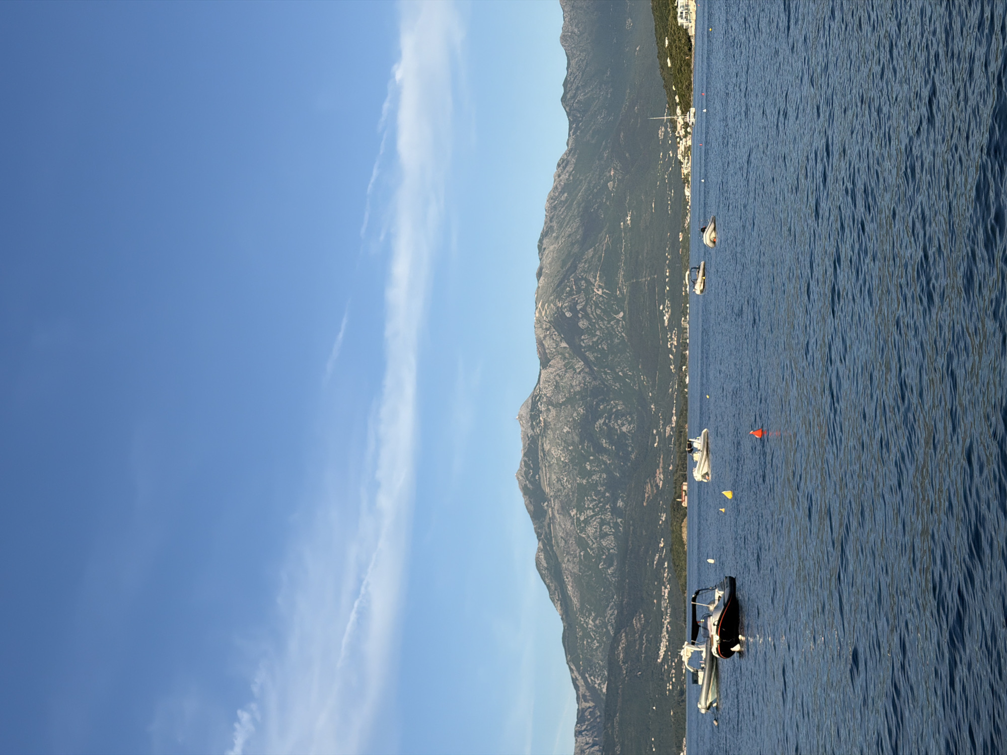 The image depicts a coastal scene, likely a part of Krašići, Montenegro. The view features a mountain range running parallel to the coastline. Clear blue sky is partially covered with long, thin clouds. The sea is calm with visible ripples and the color is a dark blue hue. A number of boats are anchored in the water, appearing along the coastline.