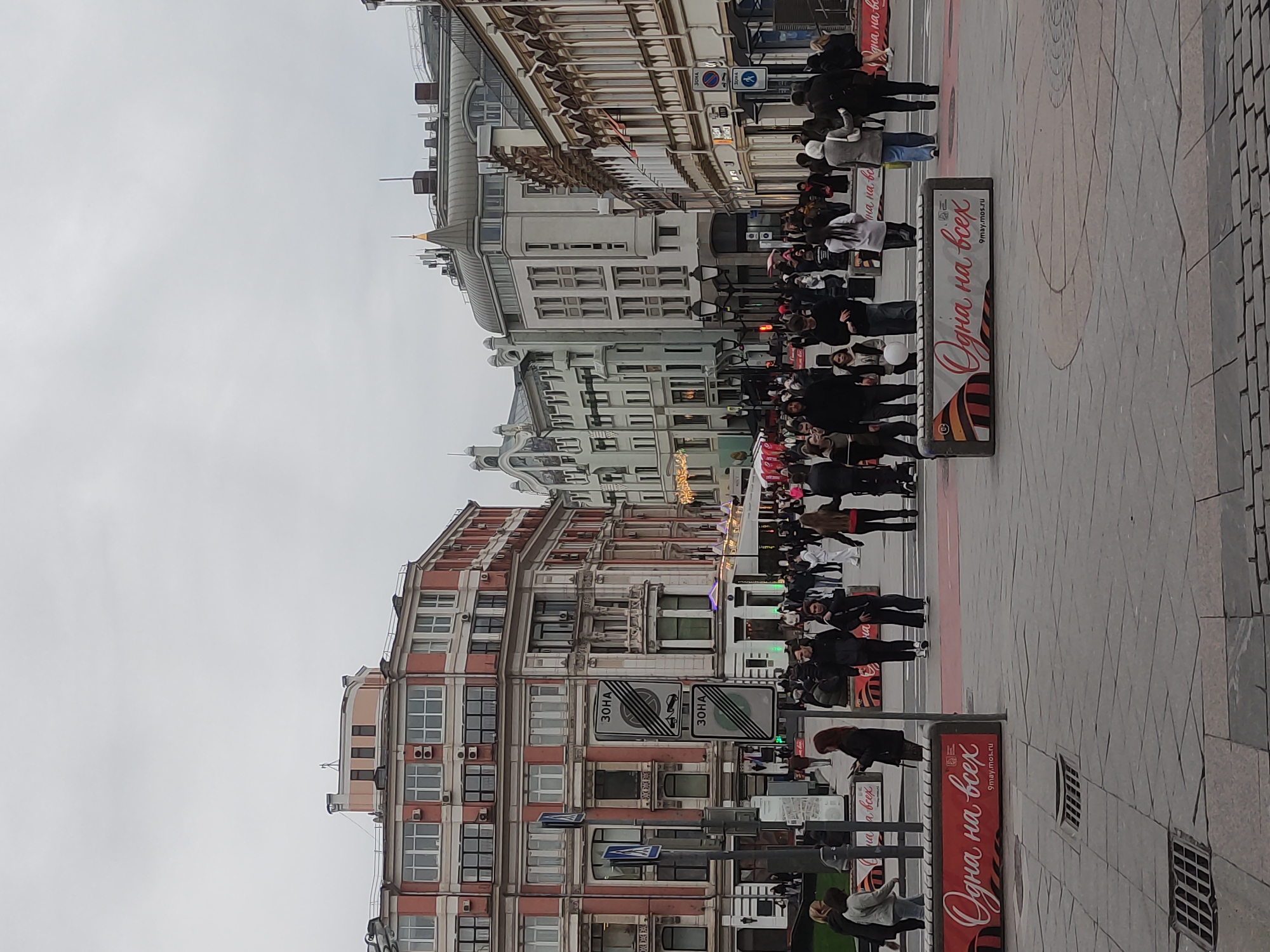 Here's a concise description of the image:
The image depicts a street in Moscow, Russia. A large crowd of people is walking along the sidewalk. Buildings line both sides of the street, featuring varied architectural styles. Street signs and advertisements are visible, including one advertising "9maymos.ru." The sky is overcast. The scene suggests a public event or gathering.