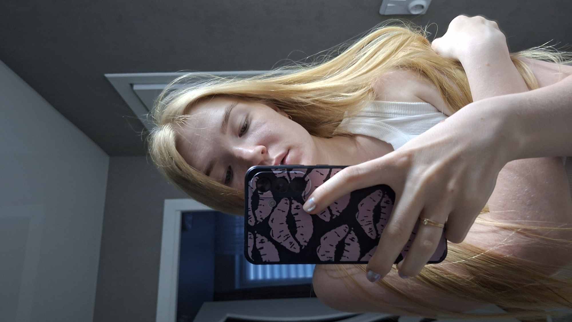 A woman with blonde hair is taking a selfie in an indoor setting, likely a bathroom, as suggested by the mirror and overhead light. She's wearing a white tank top. Her phone has a black case with pink lip prints. The background includes a door and neutral-colored walls. The location in the city of Chaikovsky, Russia is not discernible from the image.