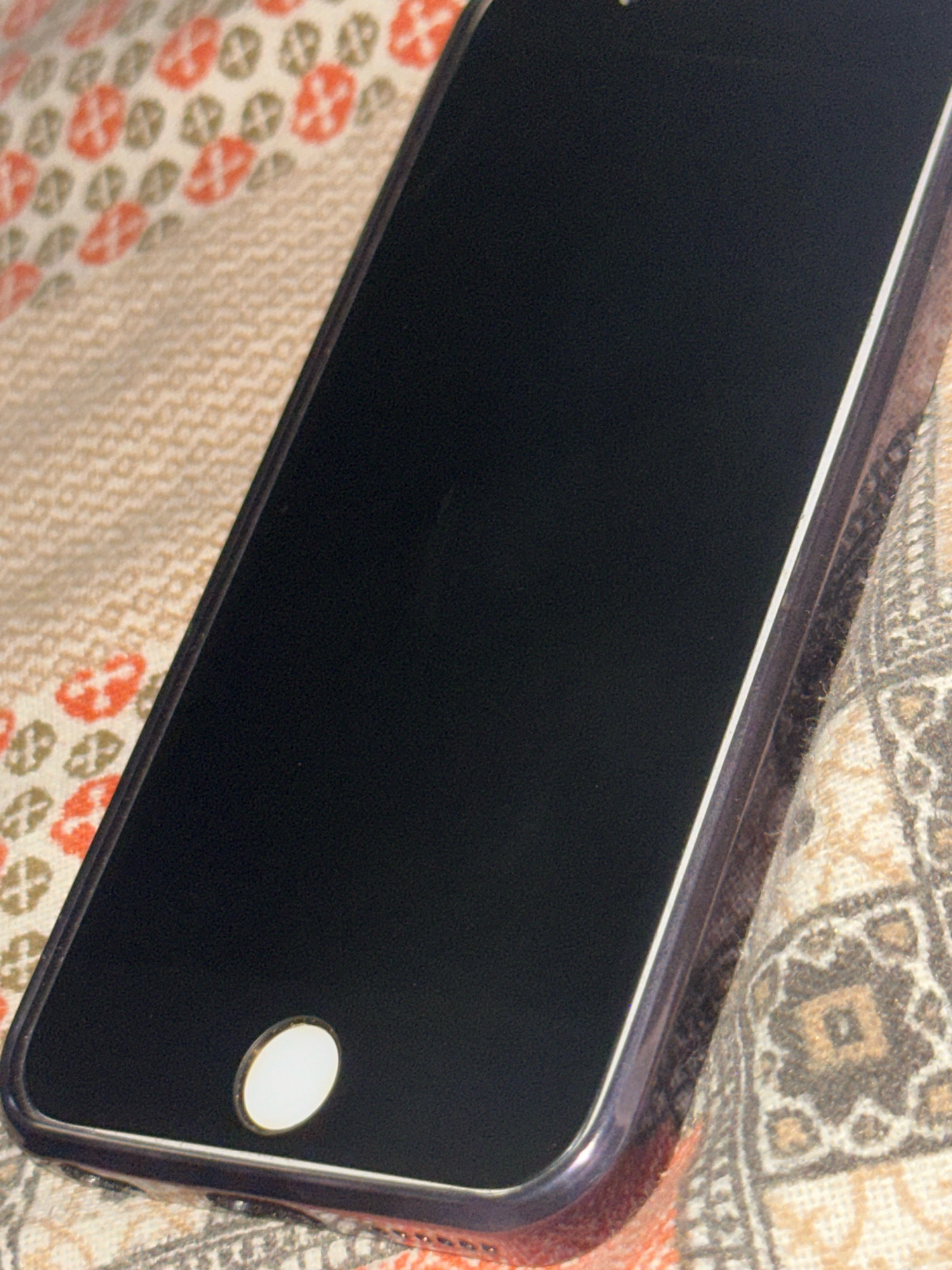 The image shows a close-up view of a black smartphone lying on a patterned fabric. The fabric appears to be a blanket or textile with a repeating pattern of orange and olive-green floral shapes on a cream background, and also a section with a geometric pattern in shades of beige and brown. The smartphone's screen is dark, indicating it is either off or displaying a dark image. The home button at the bottom of the screen is visible and illuminated. The lighting suggests an indoor environment. The location in Taharpur, India, is not discernable from the visual information provided in the image.