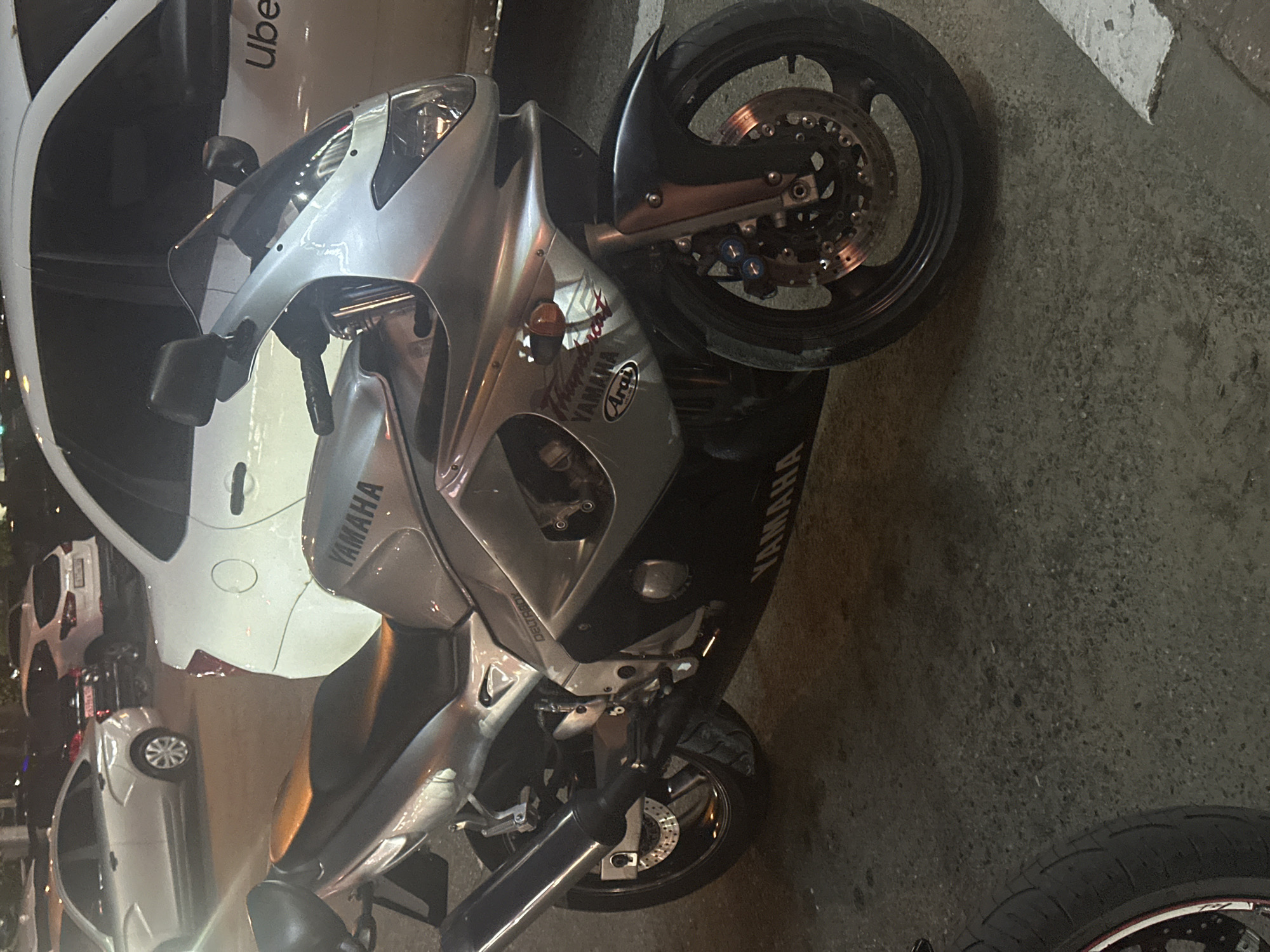 The media file presents a low-angle, sideways shot of a silver Yamaha sport motorcycle parked on what appears to be a city street or parking area at night. The motorcycle, positioned in the foreground and mid-ground, is the primary subject, displaying detailed features like its wheels, brake discs, fairings, and handlebars.
In the background and to the left of the motorcycle, several cars are visible. Most notably, a white car bears the "Uber" logo prominently on its side. Other white and darker colored vehicles are partially visible further back, suggesting a parked lineup. The scene is illuminated by artificial light sources, indicating it is nighttime. The ground is a dark, rough asphalt or concrete surface.
Notable text includes "YAMAHA" written vertically on the side fairing and horizontally on the engine casing near the front wheel of the motorcycle. A distinct "Arai" logo is also visible on the side fairing. Below the handlebar, the text "DELTABOY" can be read. The "Uber" branding on the white car in the background is clear. No people are visible in the image, and there is no specific activity taking place other than the vehicles being parked. The overall impression is that of an urban setting in Stavropol, Russia.