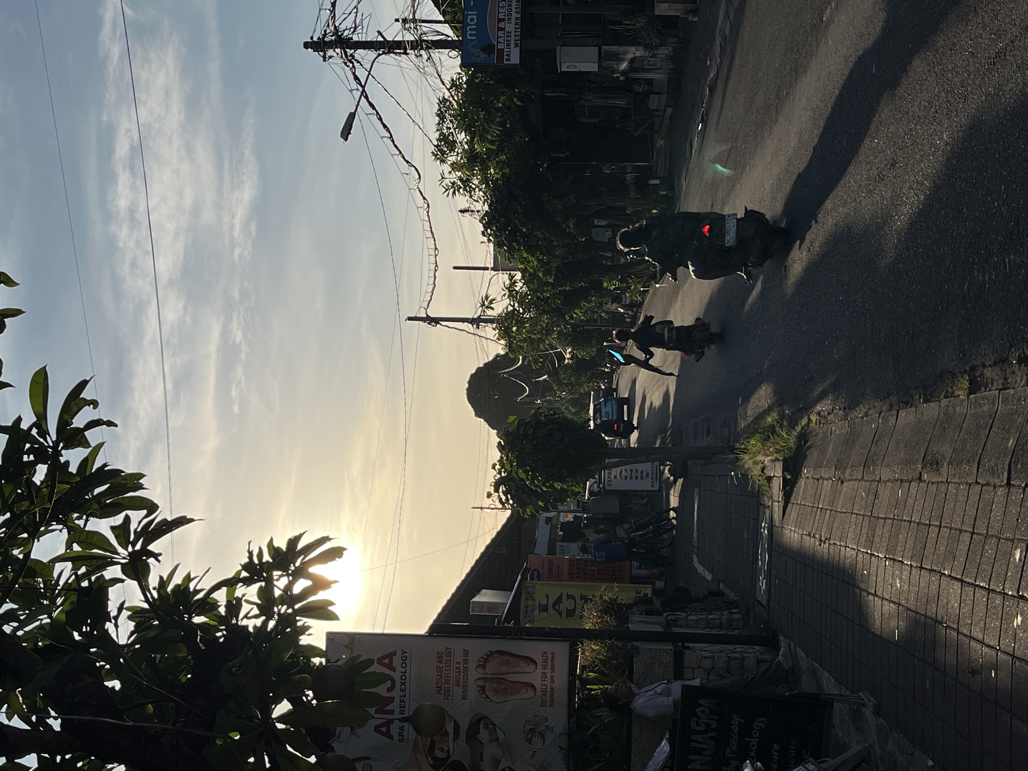 The image depicts a lively street scene in Kuta Selatan, Indonesia, bathed in the warm, golden light of either early morning or late afternoon, as suggested by the low sun casting prominent, elongated shadows across the asphalt road and sidewalk. The sky is a soft blue with scattered, wispy clouds, and the sun creates a bright glare near the horizon, partially obscured by the dark silhouettes of dense tree foliage and utility wires.
The street is a hub of activity, with multiple individuals seen on motorbikes or scooters. One person is clearly visible on a scooter, while others appear further down the road, and another figure stands or walks on the sidewalk. Overhead, a complex web of electrical wires drapes between utility poles, a characteristic feature of the setting.
The street is lined with various commercial establishments. Prominent signage includes "ANJA SPA REFLEXOLOGY," which advertises "MASSAGE AND FOOT REFLEXOLOGY" and is "SUITABLE FOR HEALTH," with text presented in both English and Russian. Other services mentioned on this sign include "Body Massage," "Reflexology," and "Manicure." Nearby, another sign points to "mai - BAR & REST," offering "BALINESE INDONESIAN WESTERN ASIA" cuisine. Additional businesses visible include a "LAUNDRY SERVICE" and a "MONEY CHANGER."
Lush green trees and bushes provide natural elements, contrasting with the man-made structures. In the background, a distant hill or natural landform rises against the sky. The combination of local commercial services, a mix of international and local languages on signs, the prevalence of motorbikes, and the general street architecture strongly indicates a Southeast Asian, specifically Indonesian, urban environment.