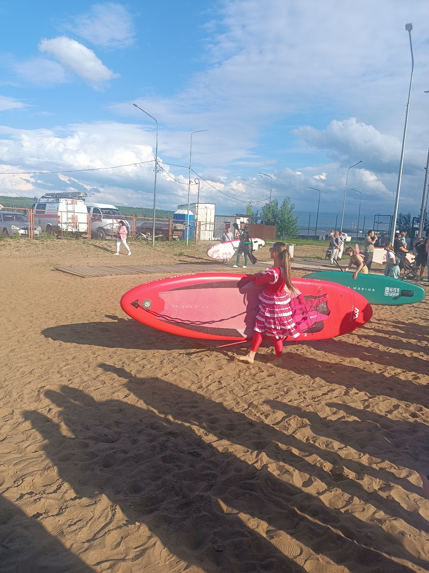 A group of people are gathered on a sandy beach, possibly near the Kama River in Chaikovsky, Russia, with paddleboards laid out on the sand. A young girl in a red and white tiered dress and red leggings stands next to a red paddleboard. Other paddleboards, one green and one white with a red pattern, are also visible. In the background, several vehicles, including vans and a car, are parked near a fence. Streetlights are spaced along the background. The sky is blue with scattered white clouds.