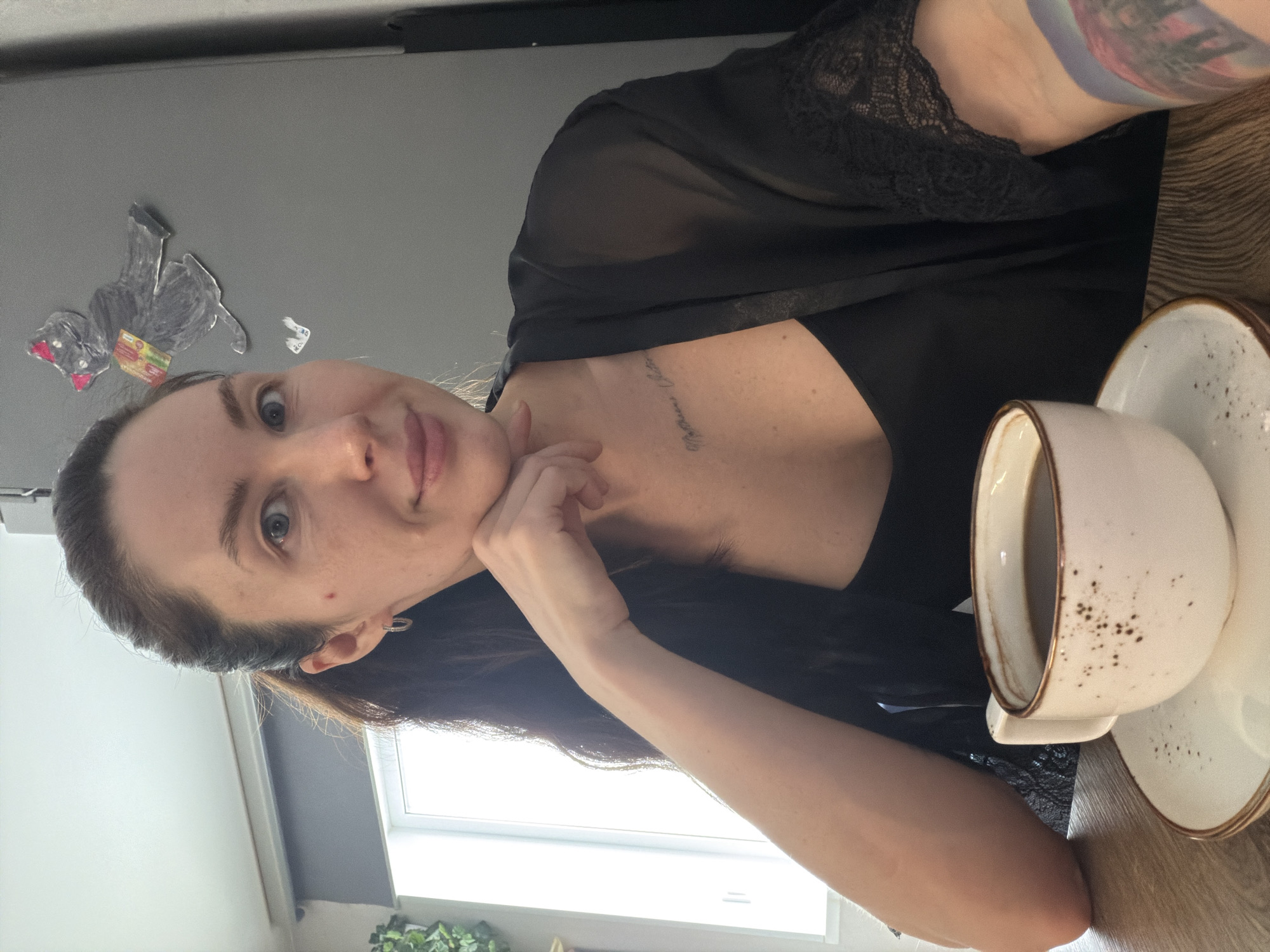 A woman with long, dark hair is seated at a table. She is wearing a black top with lace detailing. Her left arm is resting on the table, and her hand is near her chin. A coffee cup and saucer are on the table next to her. There is a tattoo visible on her chest, and another on her arm. Behind her, a window provides natural light and there is a white refrigerator on the wall. There are also drawings of a cat on the refrigerator. Based on the style of the interior, it is likely a residential space in a city in Russia. The city is Perm.