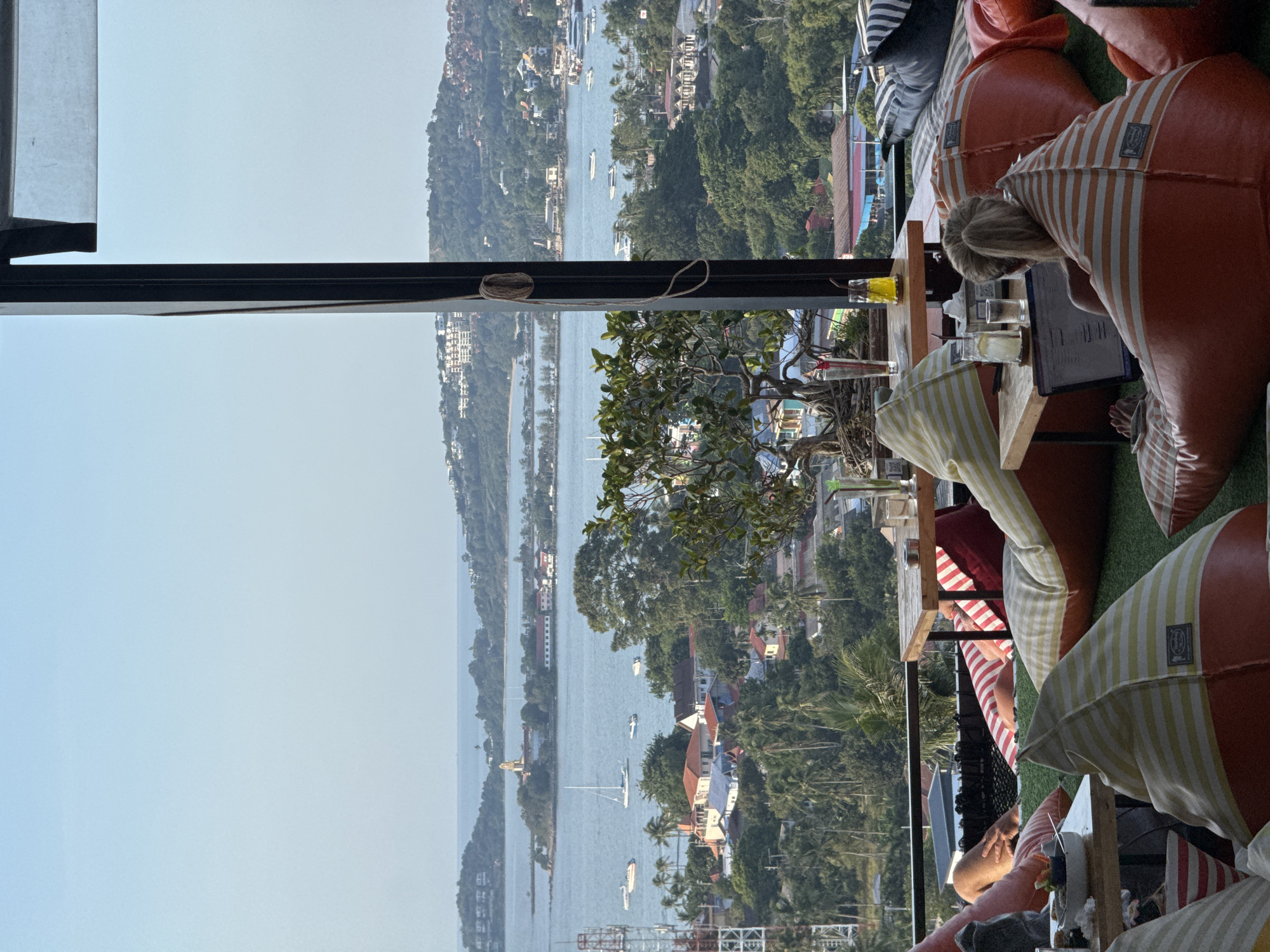 The image depicts a rooftop seating area with a panoramic view of the Ko Samui District, Thailand. In the foreground, several large, colorful beanbag chairs are arranged on a green synthetic turf. Two women are visible in the seating area. One woman, with blonde hair tied in a ponytail, is seated on an orange and white striped beanbag, facing away from the camera and working on a laptop. Another person, partially visible on the left, is reclined on a red and white striped cushion. Small wooden tables are placed between the seating areas, holding drinks and other items.
The midground reveals a lush, green hillside dotted with houses and palm trees. A body of water, likely the Gulf of Thailand, stretches into the distance, with several sailboats and smaller boats visible on its surface. In the background, hills rise, covered in vegetation and interspersed with buildings. The sky is a clear, pale blue, suggesting it is daytime. The overall scene suggests a relaxed atmosphere, possibly a cafe or restaurant with a scenic view.