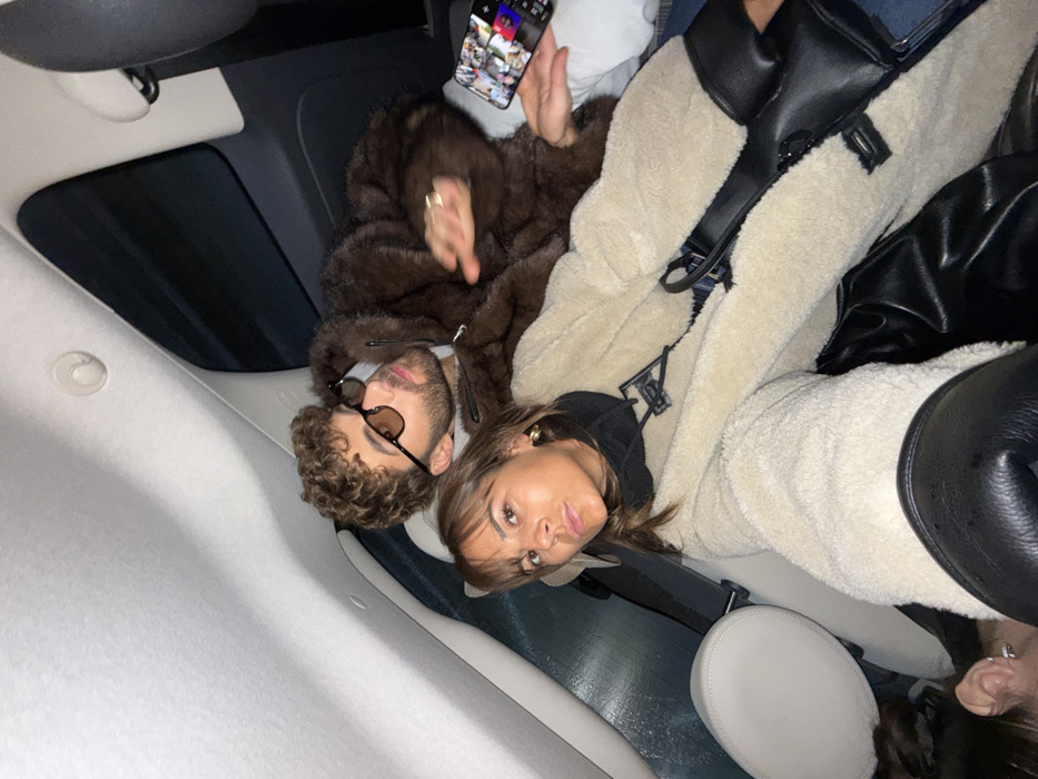 The image, taken in Villeneuve-Loubet, France, features a man and a woman inside a car, captured from a tilted perspective looking upwards. The car's light-colored ceiling and a dark sunroof are visible in the background.
On the left, a man with curly brown hair and a beard is seen wearing dark sunglasses with light frames and a brown, textured, possibly faux fur, hooded jacket. His expression is neutral. Above him, a hand holds a smartphone, which displays a grid or collage of multiple small images on its screen.
Beside the man, a woman with straight brown hair looks towards the camera. She wears a black top or hoodie underneath a cream or beige shearling-lined jacket. A black stylized logo, resembling an 'LV' or 'V', is prominently visible on the front of her jacket. She has an earring in her right ear and her expression is somewhat serious or contemplative. A black bag or strap is draped over her shoulder. Part of another person's head and hair can be seen in the bottom right corner of the frame. The subdued lighting suggests an indoor setting, possibly evening or night.