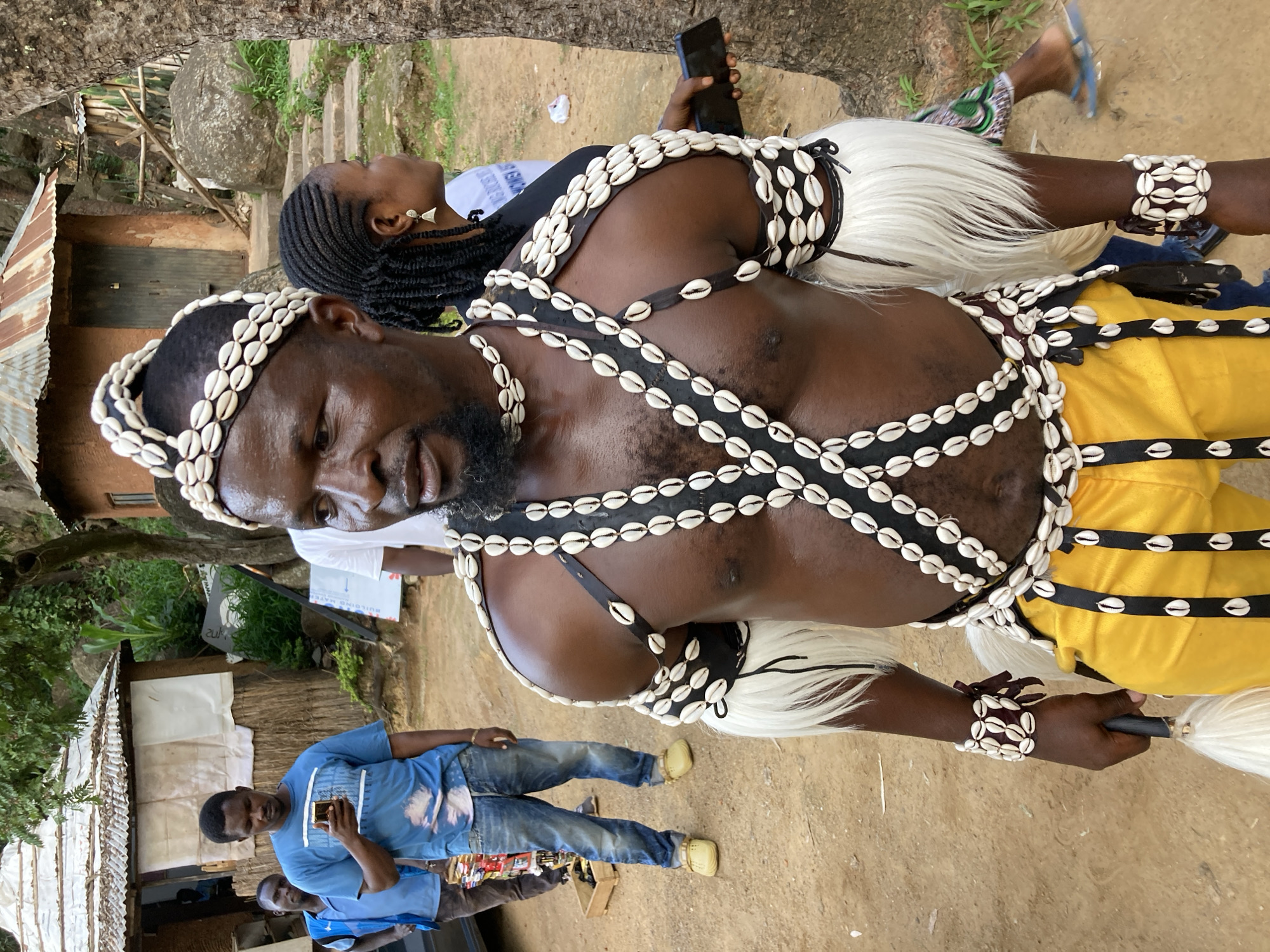 In Jos, Nigeria, a man adorned in traditional attire, featuring a headdress, necklaces, and armbands made of cowrie shells, stands in the foreground. He wears a sleeveless black garment across his chest and arms, embellished with multiple rows of cowrie shells. His lower body is clad in vibrant yellow trousers. The man holds a whisk-like object in his left hand. Behind him, other individuals are visible, including a man in a blue t-shirt and jeans, looking at his phone, and a woman with braided hair. The setting appears to be a rural or semi-rural environment, with rustic huts and a large tree in the background. The ground is sandy and dirt.