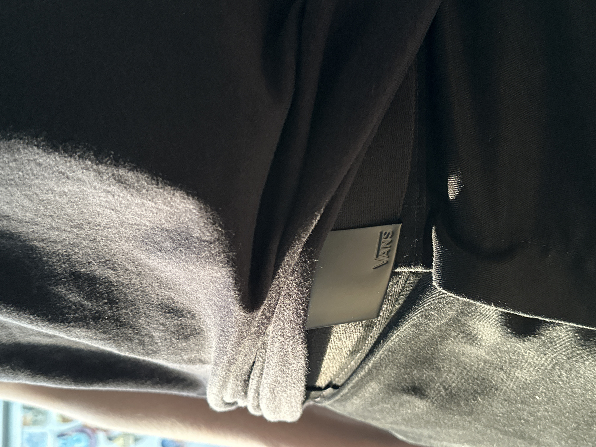 The image shows a close-up of a person wearing black jeans and a dark gray t-shirt. A black belt with a silver buckle that has the word "VANS" embossed on it is visible. The t-shirt is tucked into the jeans and cinched around the waist, creating folds. Sunlight illuminates the scene from the right, casting shadows and highlighting the textures of the fabric. The background is out of focus and appears to be a wall with some colorful items, possibly posters or artwork. There is no indication of the location being in Moscow, Russia.