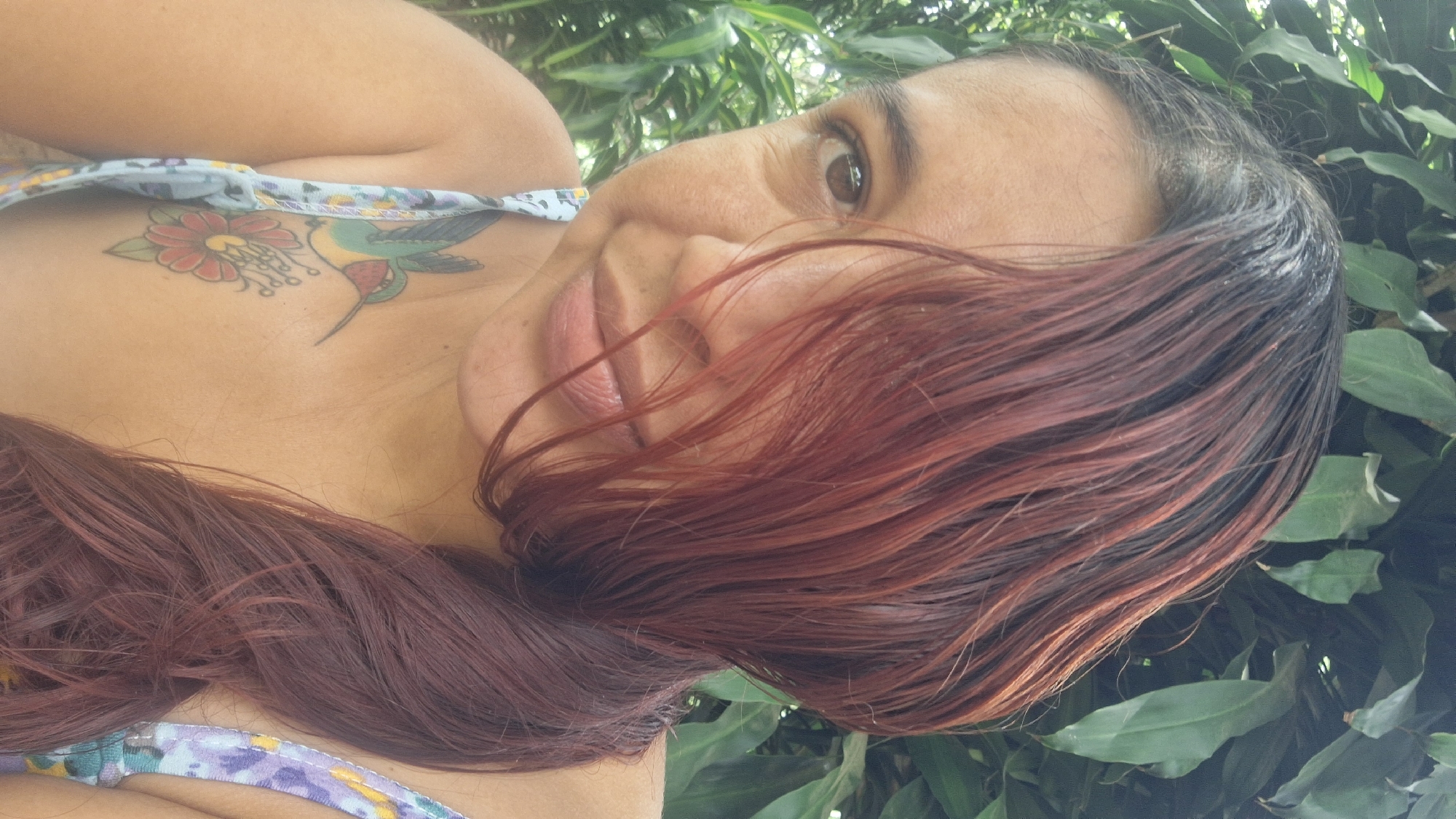 A close-up portrait, oriented diagonally, features a woman outdoors. Her head is tilted, with her right eye looking towards the camera and a gentle smile on her lips. Long, dark red hair, appearing somewhat wet or styled, flows across her face and chest, with darker roots visible near her scalp.
She is wearing a light-colored top with a patterned design, visible on her shoulder strap and upper chest. A prominent tattoo is inked on her upper right chest, depicting a detailed red and yellow flower, possibly a fuchsia, alongside a colorful hummingbird with green, blue, and yellow plumage.
The background consists of abundant, lush green foliage, suggesting a natural outdoor setting, likely a garden or tropical environment. The scene is brightly illuminated by natural daylight. The woman's expression is calm and pleasant. The setting, as provided, is Vallecito, Venezuela. No visible text is present in the image.