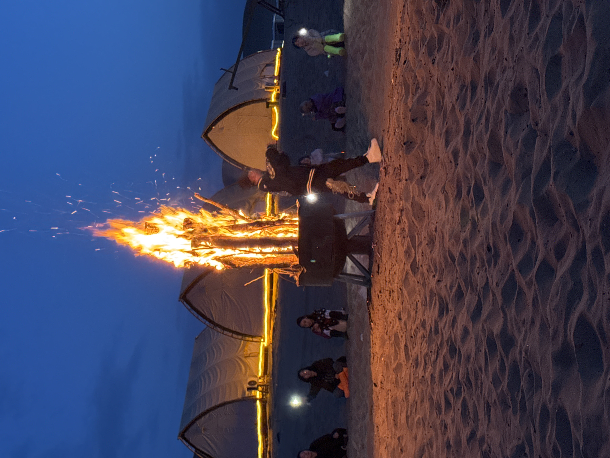 The image captures a nighttime scene on a sandy terrain, possibly a desert or beach in Dunhuang City, China, given the subject and context. A large bonfire is the focal point, with flames reaching high and casting light on the immediate surroundings. Several people are gathered around the fire, some seated or crouching. In the background, there are tent-like structures, possibly part of a campsite, illuminated by lights. The sky is a deep blue, indicating it is either dusk or night. One person is standing near the fire, with their back to the camera, wearing what appears to be casual clothing.