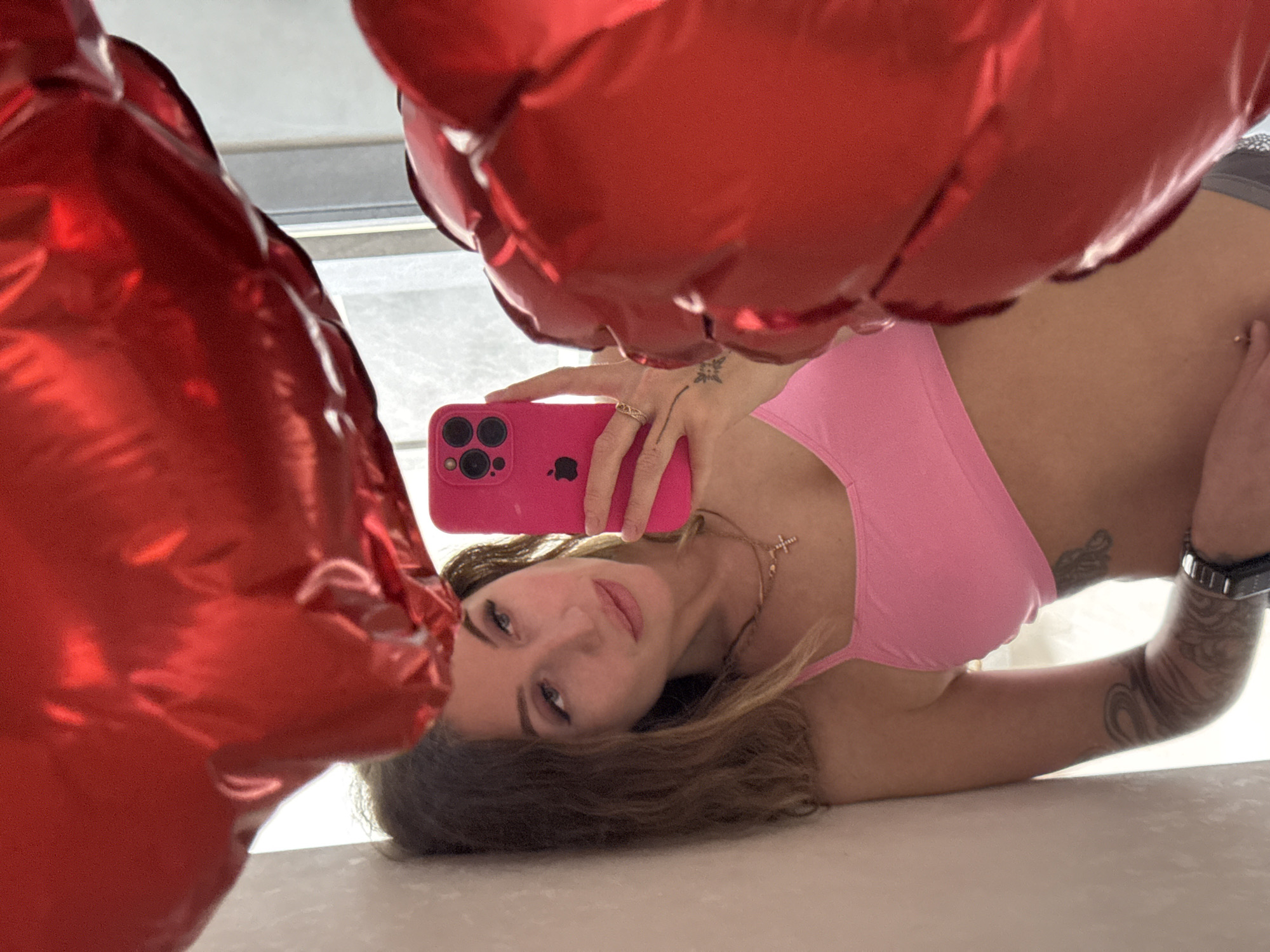 A mirror selfie captures a young woman lying on a light-colored surface, reflected upside down in the frame. She has long brown hair fanned out around her head and looks directly at the camera with a neutral to slightly smiling expression. She wears a pink sports bra or top, a necklace with a cross pendant, and a watch on her left wrist. Tattoos are visible on both of her arms and a small star-like tattoo is on the back of her right hand.
She holds a bright pink iPhone with a multi-lens camera system in her right hand, taking the photograph. A gold-colored ring is on her right-hand ring finger. Large, glossy red metallic balloons, possibly heart-shaped, dominate the foreground, framing her reflection and adding a festive or romantic element to the scene. The background, visible behind the balloons and through the reflection, suggests an indoor setting with natural light, possibly from a window. The lighting indicates it is daytime. The partial Apple logo is visible on the phone case. The overall composition creates an intimate and playful atmosphere. No specific visual cues in the image confirm the location as Chaikovsky, Russia.