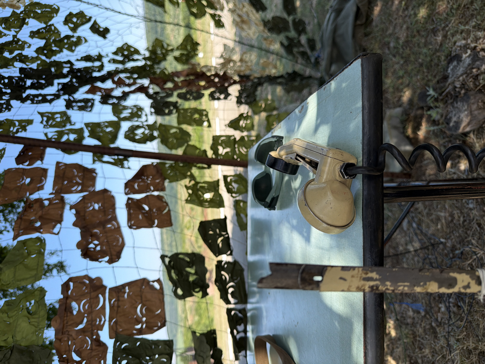 The image depicts an outdoor scene with a table in the foreground, likely in Beliakovets, Bulgaria. On the table, there is a vintage-style telephone receiver with a coiled cord. To the left of the receiver, a portion of a strap or belt is visible, along with a metal pole with chipped paint. In the background, a camouflage netting is hung on a fence, creating a dappled effect against a bright sky and a green, hilly landscape. The ground appears to be covered with dry grass, dirt, and some scattered wires. The overall impression is one of a temporary or rustic setup, possibly related to a historical reenactment or a field observation post.