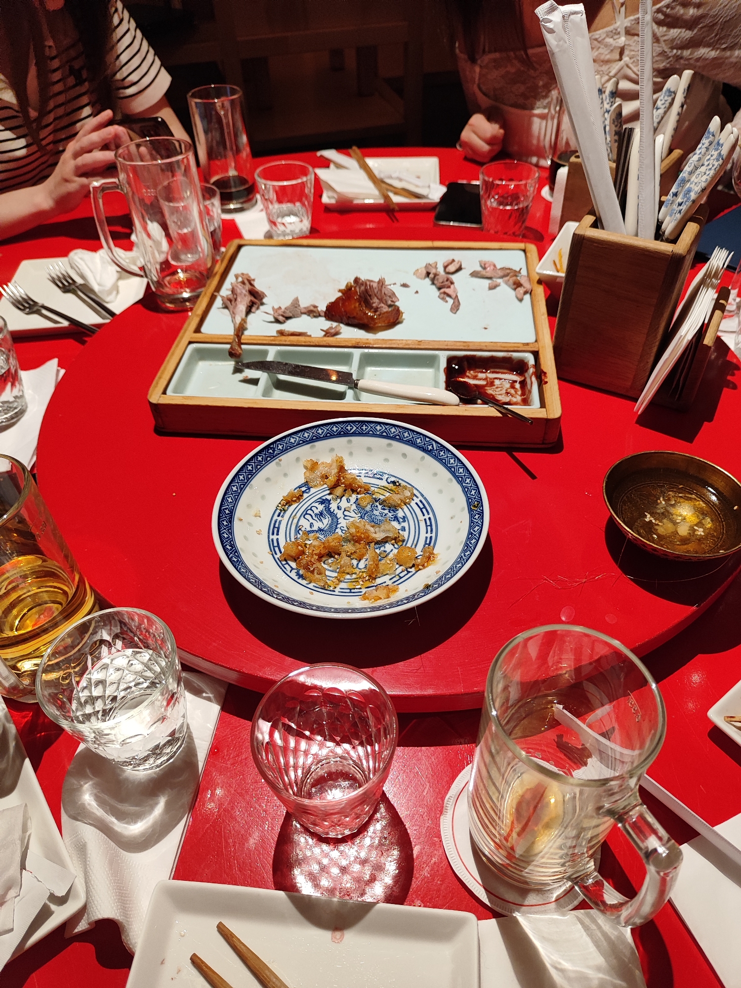 The image captures an overhead view of a red dining table, likely in an East Asian restaurant, after a meal. The circular table has a lighter red rotating center.
At least two individuals are partially visible at the table. On the left, a person wearing a black and white striped top is seen, with their hands near a smartphone. On the right, another person in a light-colored, possibly lace, top is partially visible.
The table is set with various items. A light blue rectangular serving tray in a wooden frame holds leftover meat, appearing to be roasted poultry like duck, along with some bones and dark sauce in partitioned sections. A knife is also on this tray. A blue and white patterned plate in the foreground contains scattered crispy, golden-brown fried bits, possibly from the poultry skin or a garnish. A small brass or golden bowl next to it holds a liquid, possibly oil or a light broth, with white sesame seeds on top.
Numerous clear glasses are spread across the table: some empty, some holding water or a light-colored beverage, and one large beer mug with a handle. Several white square plates, some with forks and knives, and white napkins are also present. A wooden holder contains multiple white-wrapped chopsticks or straws, along with decorative ceramic chopsticks.
The scene suggests the aftermath of a shared meal. The artificial lighting indicates an indoor setting, likely evening. There is no discernible text visible in the image.