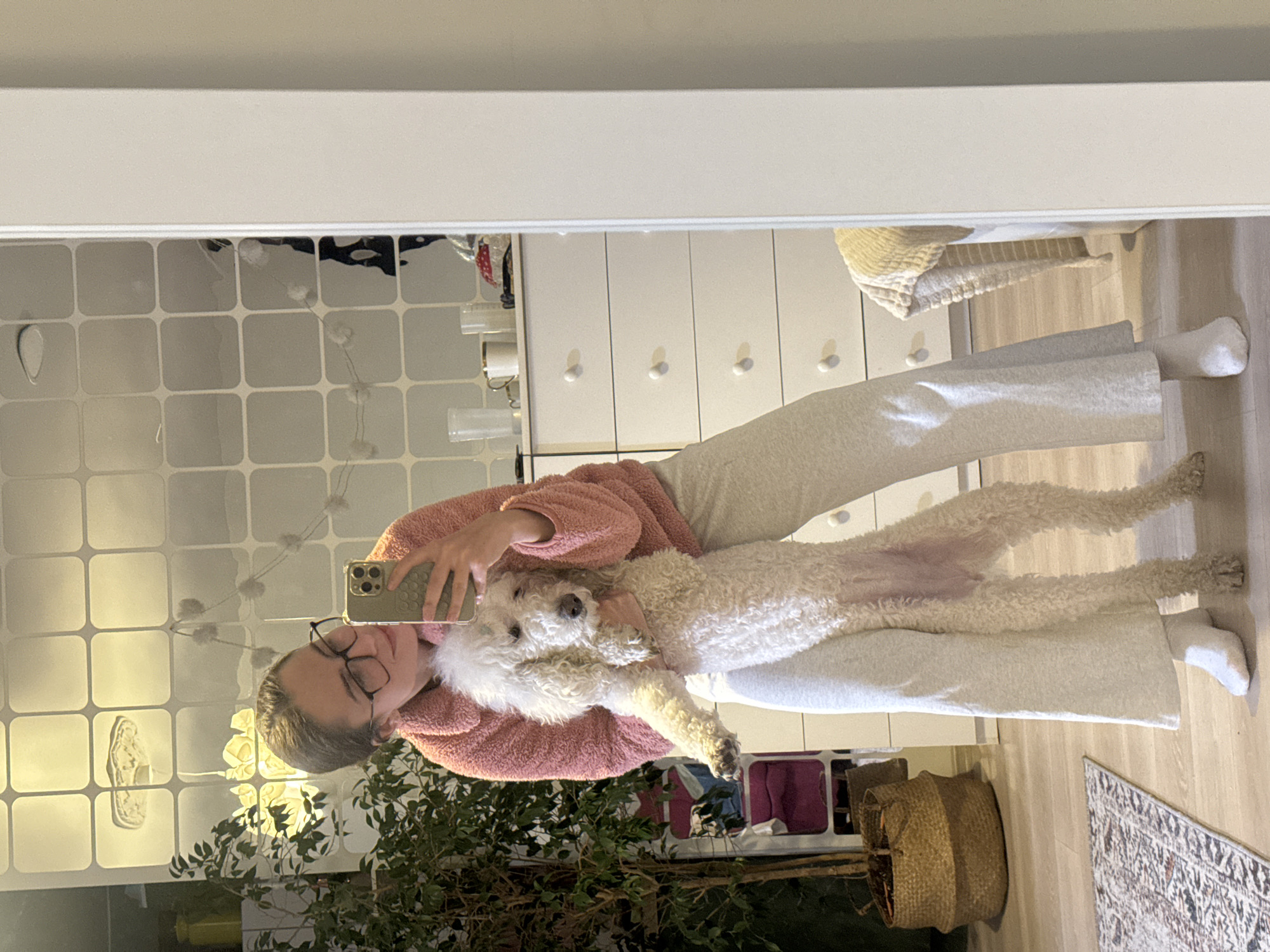 A fair-skinned person wearing glasses, a pink fuzzy top, and light-colored sweatpants and socks is taking a selfie in a mirror while holding a cream-colored standard poodle dog. The background includes a white dresser, a tiled wall covering, plants in a basket, and a rug. The location cannot be identified as Krasnoyarsk, Russia from the given image.