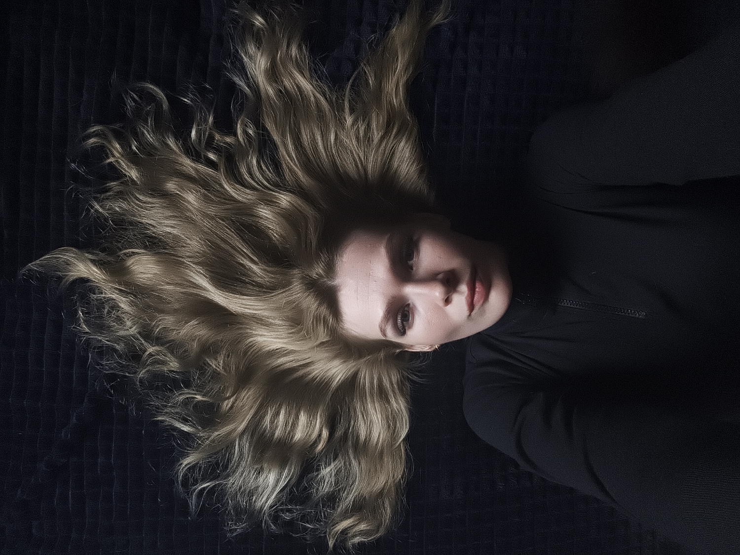 The image captures a young woman lying on a dark, ribbed, textured surface, likely a blanket or upholstered fabric. She is positioned diagonally across the frame, with her head oriented towards the upper left and her body extending towards the lower right.
The woman has long, wavy, blonde or light brown hair that is fanned out around her head, creating a halo-like effect against the dark background. Her face is illuminated by soft, directional light, highlighting her features. Her eyes are open, gazing directly towards the camera, and her lips are slightly parted, revealing a subtle, neutral, and perhaps pensive expression. She wears a dark, long-sleeved top, possibly a turtleneck or athletic wear, with a visible zipper detail around the collar area. The majority of her body and clothing are submerged in shadow, creating a strong contrast with her illuminated face and hair.
The scene suggests an indoor setting, likely a relaxed environment such as a bedroom or living space. The lighting conditions appear to be low, contributing to a dramatic and intimate mood. There are no explicit visual cues within the image to indicate a specific time of day or weather conditions. No text is visible in the media file. Given the provided context, the location of this scene is Русский Вожой, Russia.