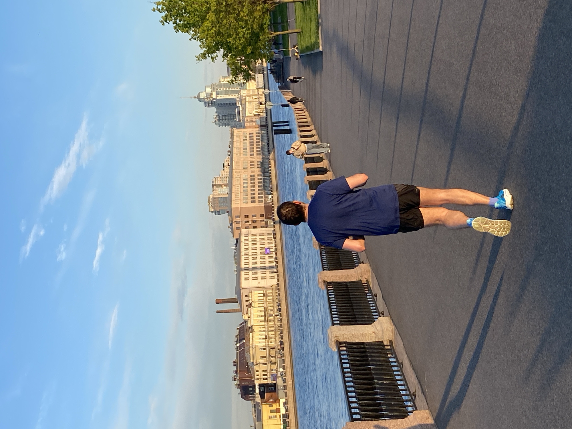 Here's a detailed description of the image:
**Scene:** An outdoor urban environment in Saint Petersburg, Russia. A river runs through the city, with buildings lining the embankment. The sky is blue with light clouds.
**People:** A man is the main subject, seen from the back, wearing a dark blue t-shirt, black shorts, and running shoes with blue accents. He is positioned on the embankment next to the river. Other people are walking along the embankment in the distance.
**Objects:** Key objects include the river, the buildings along the embankment (various architectural styles and colors), the embankment itself with metal railings, and trees.
**Actions/Interactions:** The man appears to be exercising or running. The other people are walking.
**Environment:** The setting is Saint Petersburg, evident from the architecture and riverfront. It is daytime, with a clear sky suggesting fair weather. The location appears to be a popular spot for recreation and walking.