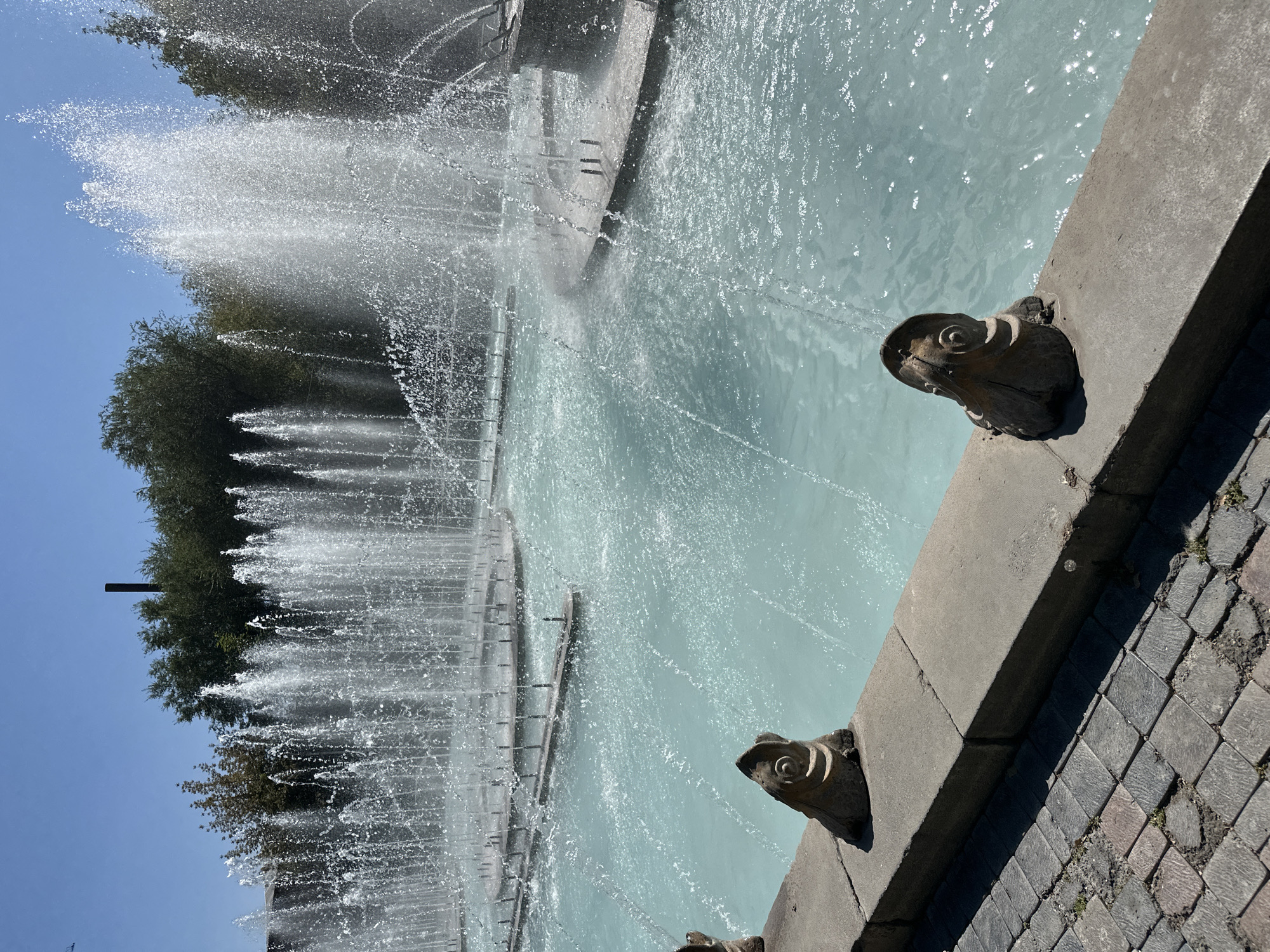 The image depicts a large, multi-tiered fountain with numerous jets of water spraying upwards and outwards, creating a misty effect against a clear blue sky. The water in the fountain basin is a light, turquoise blue. In the foreground, a stone ledge runs along the edge of the fountain. Two decorative stone elements resembling stylized frogs are perched on this ledge, facing towards the fountain. To the left of the ledge, a cobblestone pathway is visible. Behind the fountain, a line of lush green trees forms a backdrop, with a dark, cylindrical structure, possibly a chimney or tower, rising above the trees in the distance. The overall scene suggests a public outdoor space on a sunny day. This fountain is likely part of the Cascade complex in Yerevan, Armenia, known for its elaborate fountains and outdoor art installations.