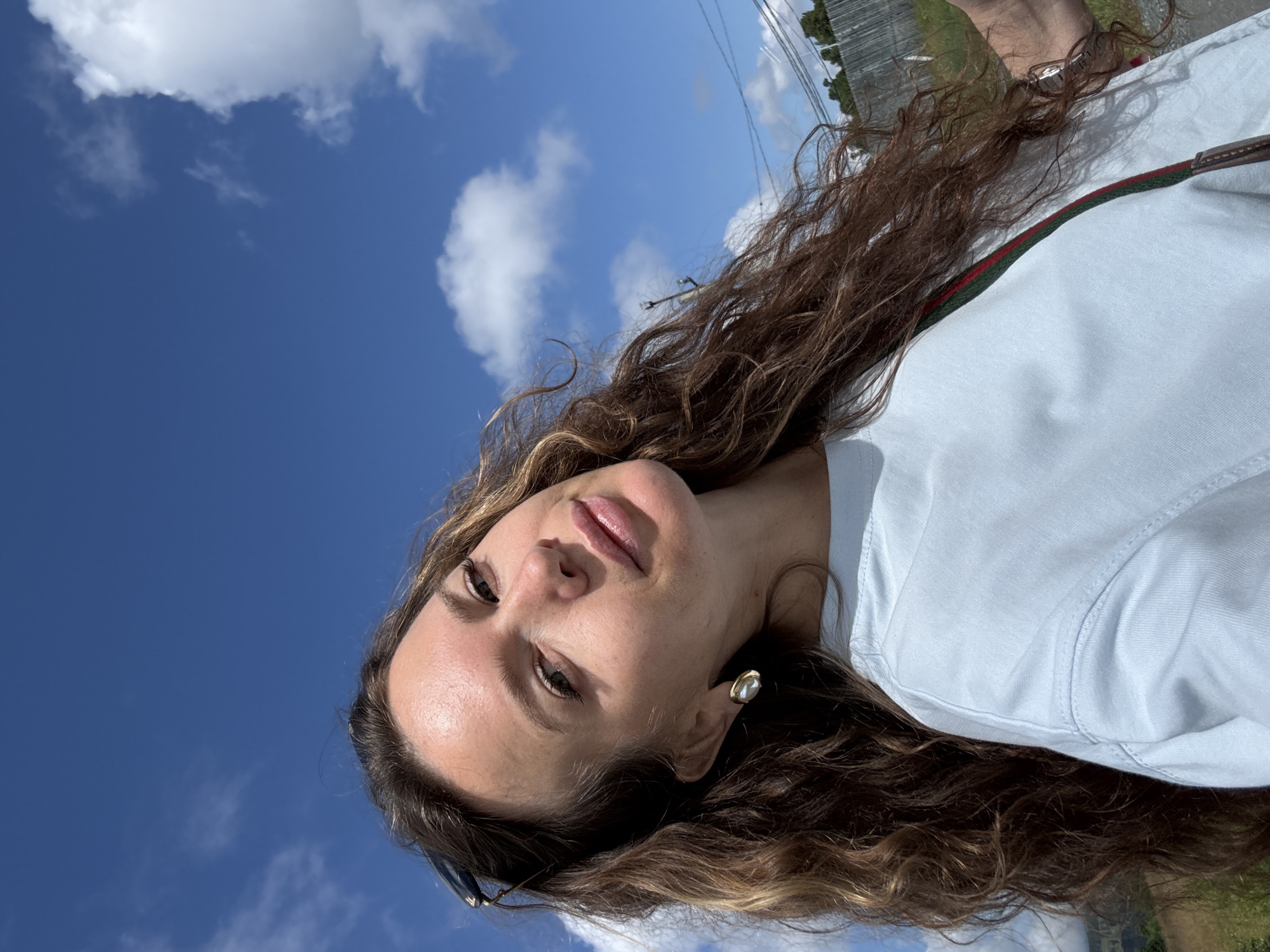 A medium shot selfie shows a woman with long, curly brown hair, wearing a light blue t-shirt. She is outdoors under a blue sky with scattered white clouds. She is wearing a single gold earring with a pearl-like stone. Sunglasses are perched on top of her head. The background shows a fence and some trees. The image is in focus and well-lit. The exact location within T/B Seminskii Pereval, Russia is not discernible from the image.