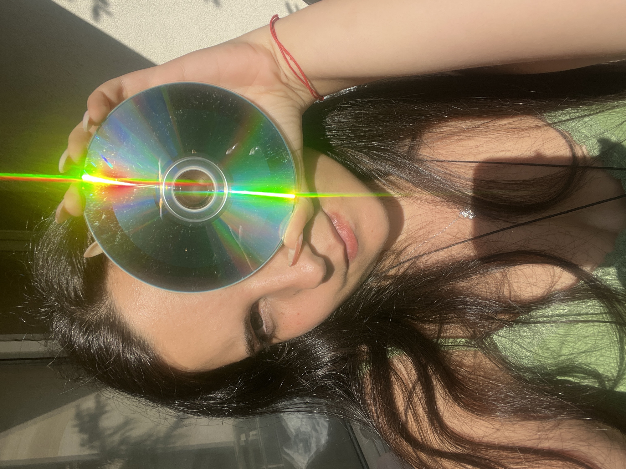 A medium shot captures a young woman holding a CD in front of her face, obscuring her left eye and part of her nose. She has long, dark hair and wears a green top. The CD reflects rainbow light, creating a prism effect. Sunlight casts shadows on the wall behind her. The scene appears to be indoors or on a balcony, with natural light suggesting daytime. There are no other people or animals visible. The location is not specified in the image.