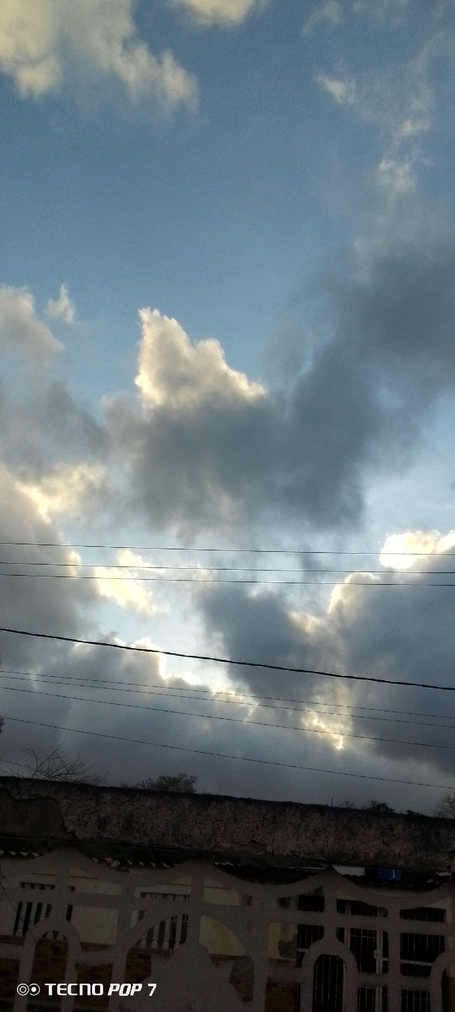 This is a low-angle photograph taken from Altagracia, Venezuela, capturing a cloudy sky with beams of sunlight breaking through. The composition is dominated by the sky, which fills approximately two-thirds of the frame. Dark gray and white clouds are visible, with patches of bright sunlight illuminating them from behind. Several black power lines are strung horizontally across the middle of the image, intersecting the cloudy sky. Below the power lines, the upper portion of a low-rise building is visible, featuring a textured, light-colored facade with some areas of peeling paint. A white decorative fence or wall with an ornate design runs along the bottom of the building. The scene appears to be outdoors, likely during daylight hours given the presence of sunlight. A watermark in the bottom left corner reads "TECNO POP 7".
